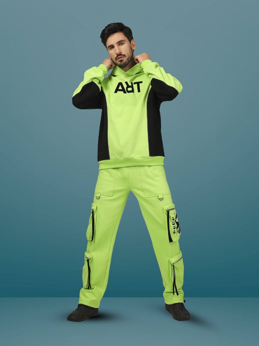 Model wearing fluorescent multi-pocket cargo pants in neon green, combined with a matching hoodie on a blue background.