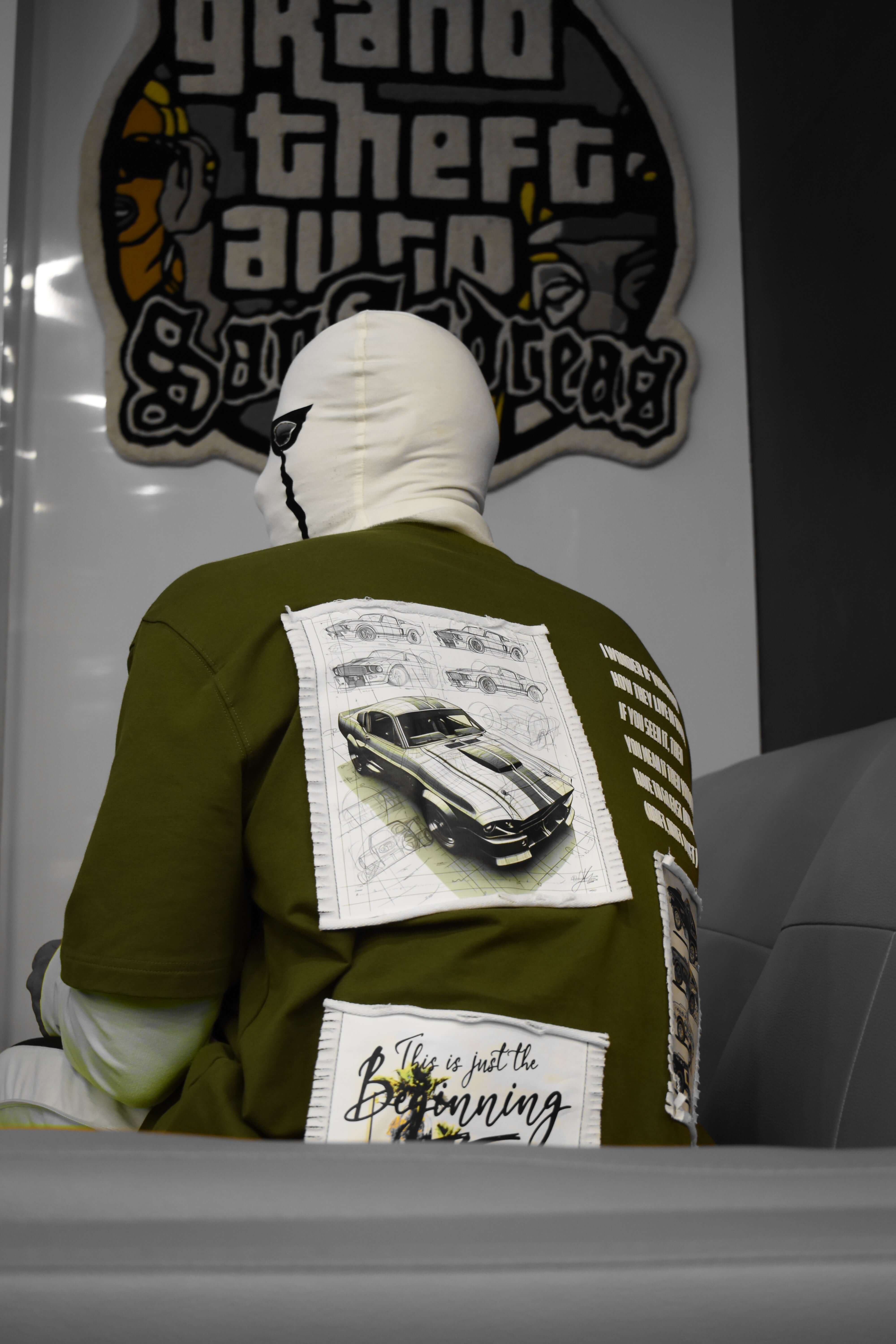 Rear view of person wearing an oversized olive green Ford Mustang t-shirt with car design, in a gaming-themed room.