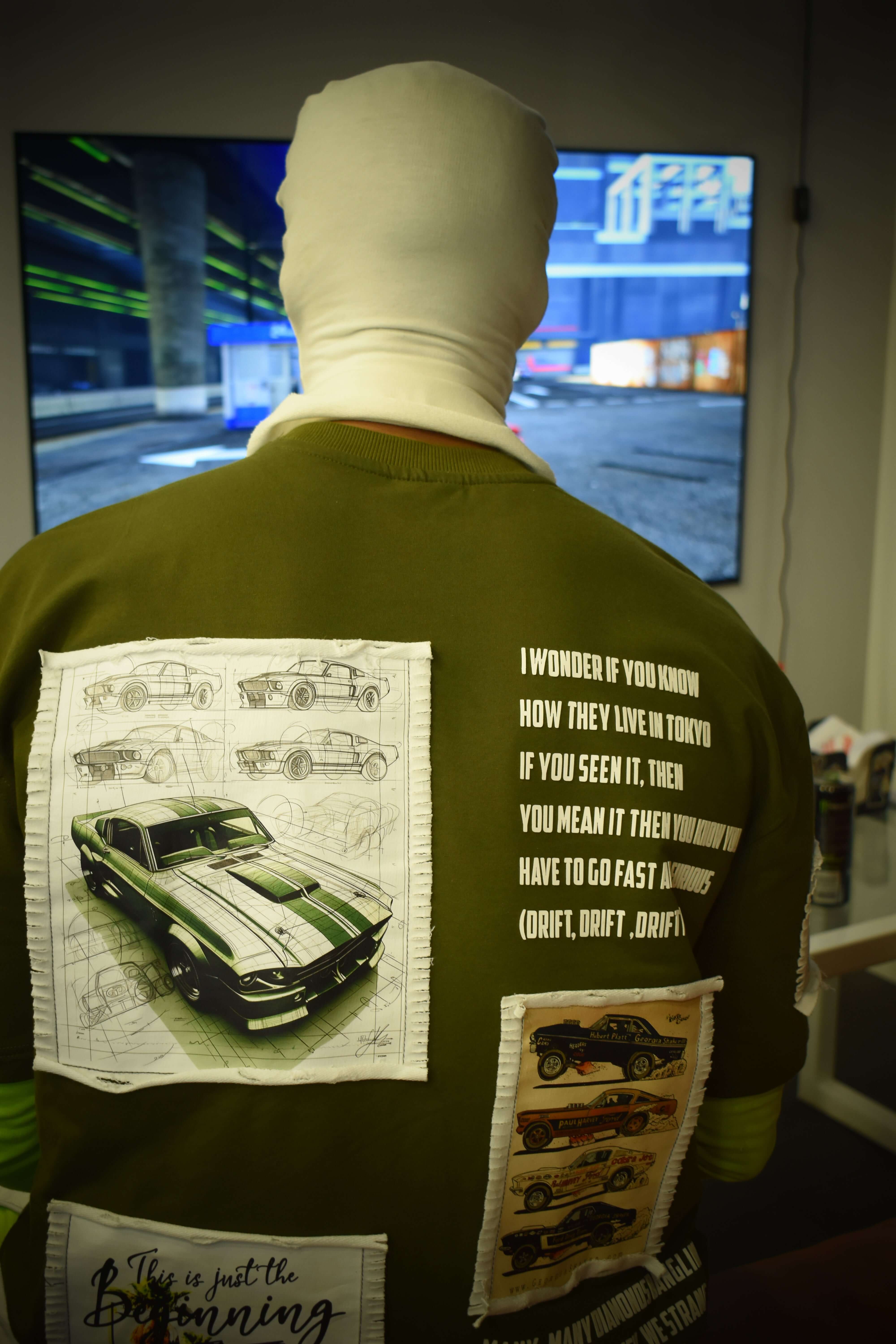 Person wearing olive green oversized Ford Mustang designer T-shirt with car graphics and text, standing indoors.
