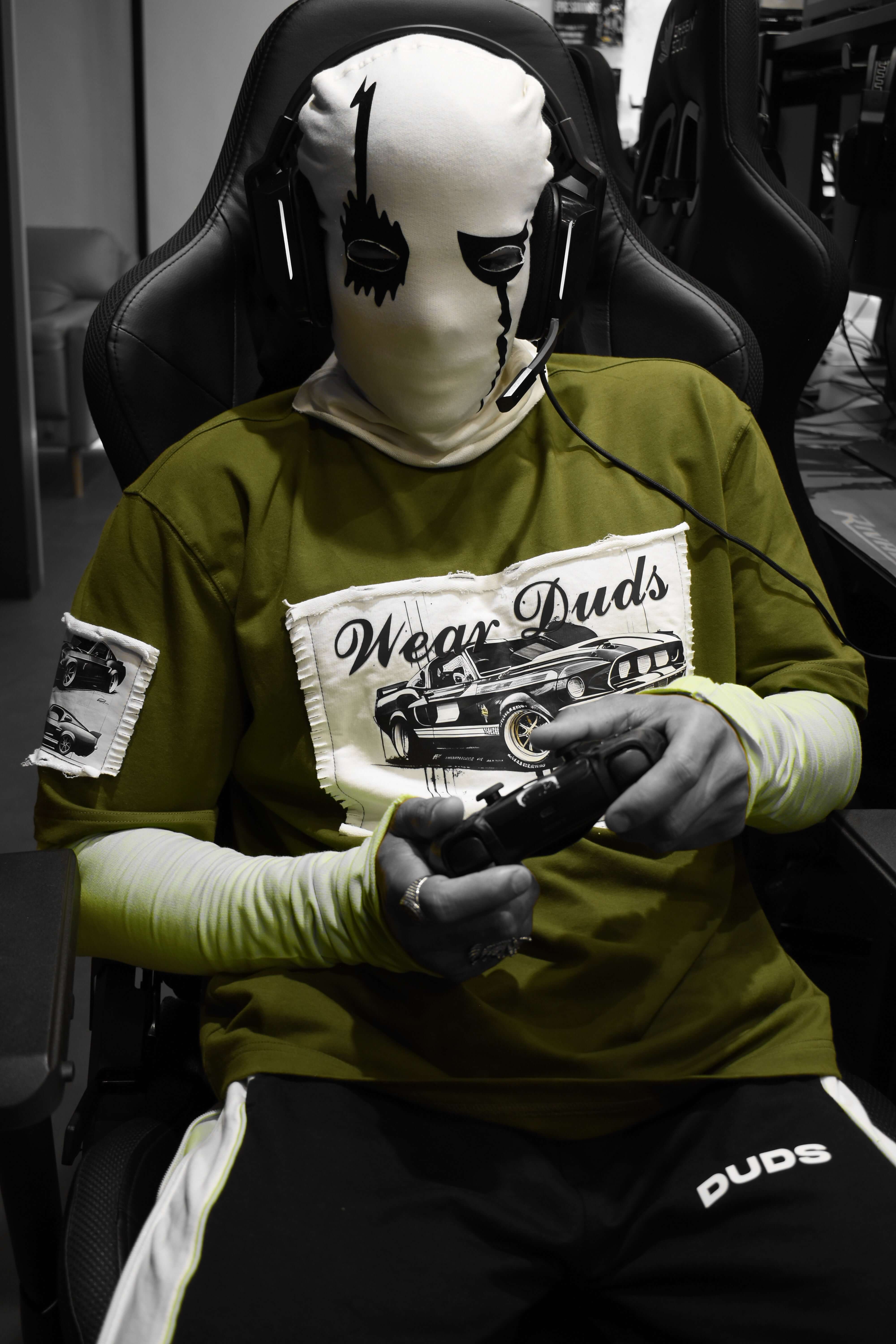Person wearing olive green oversized t-shirt with vintage car graphic, gaming in chair, headphones on; Ford Mustang style; stylish gamer look.