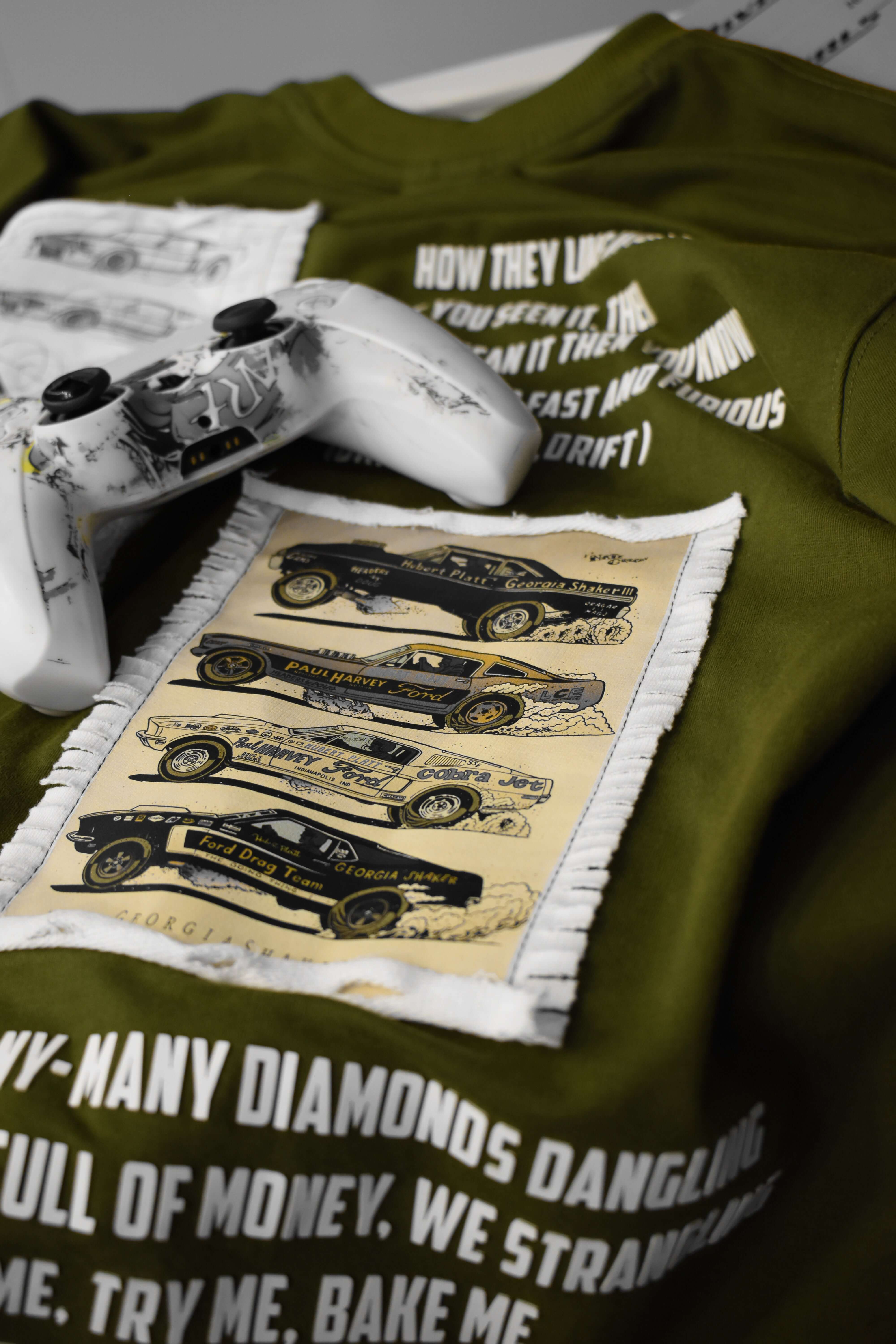 Olive green Ford Mustang designer t-shirt with car graphics and white gaming controller on textured fabric.
