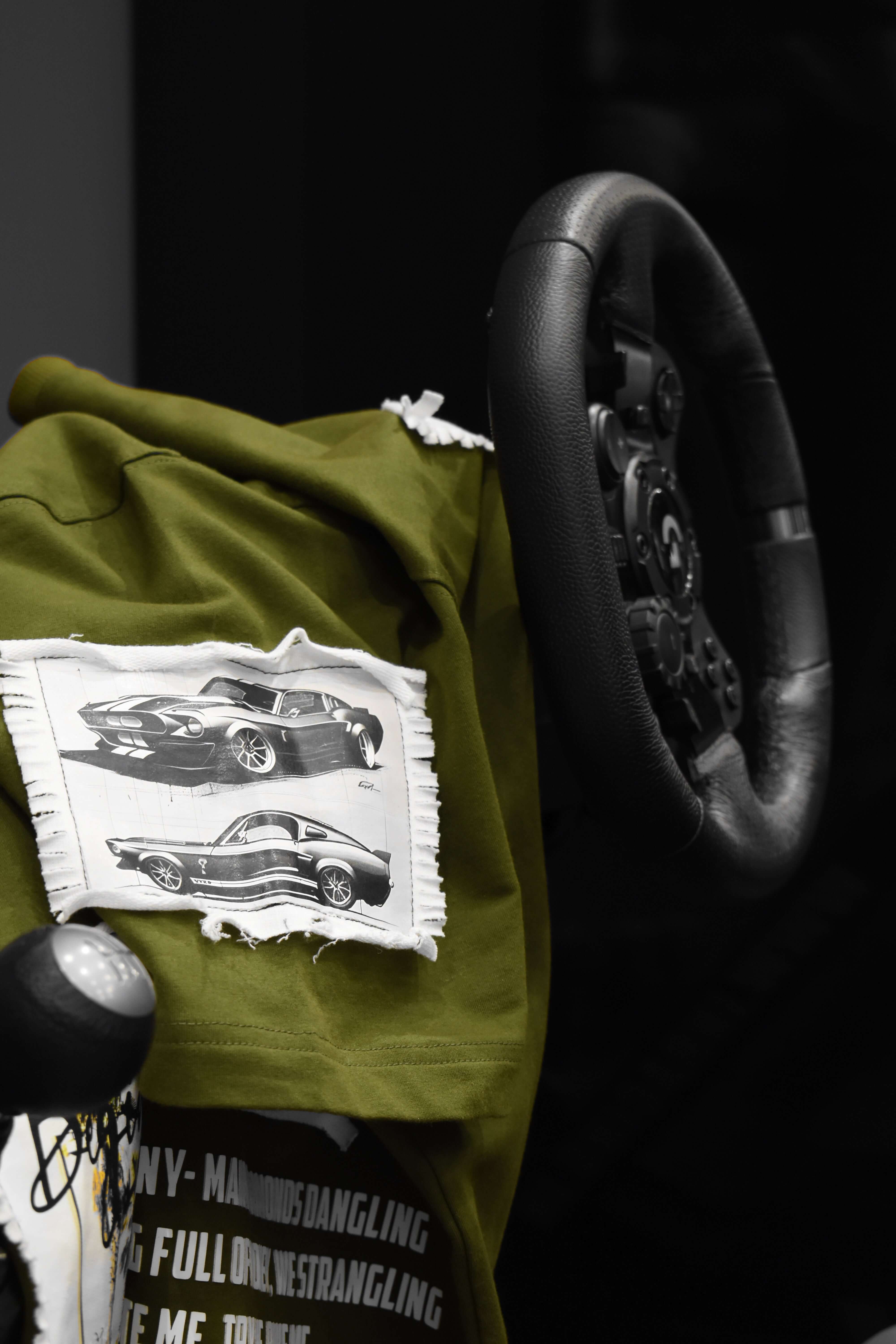 Olive green Ford Mustang oversized t-shirt draped over a steering wheel, showcasing vintage car design patches.