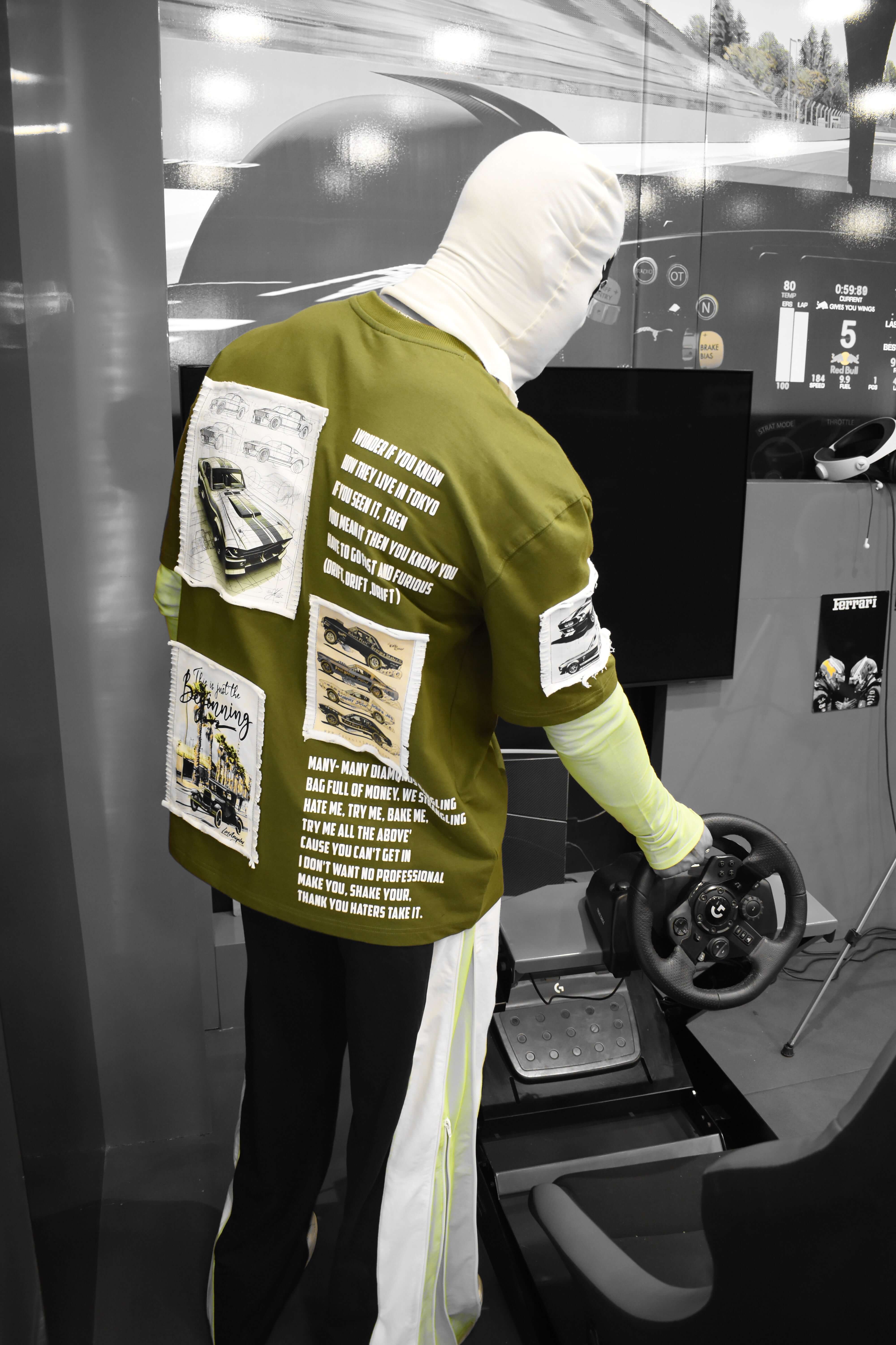 Person wearing Ford Mustang olive green oversized T-shirt with print, holding a steering wheel in a modern setting.