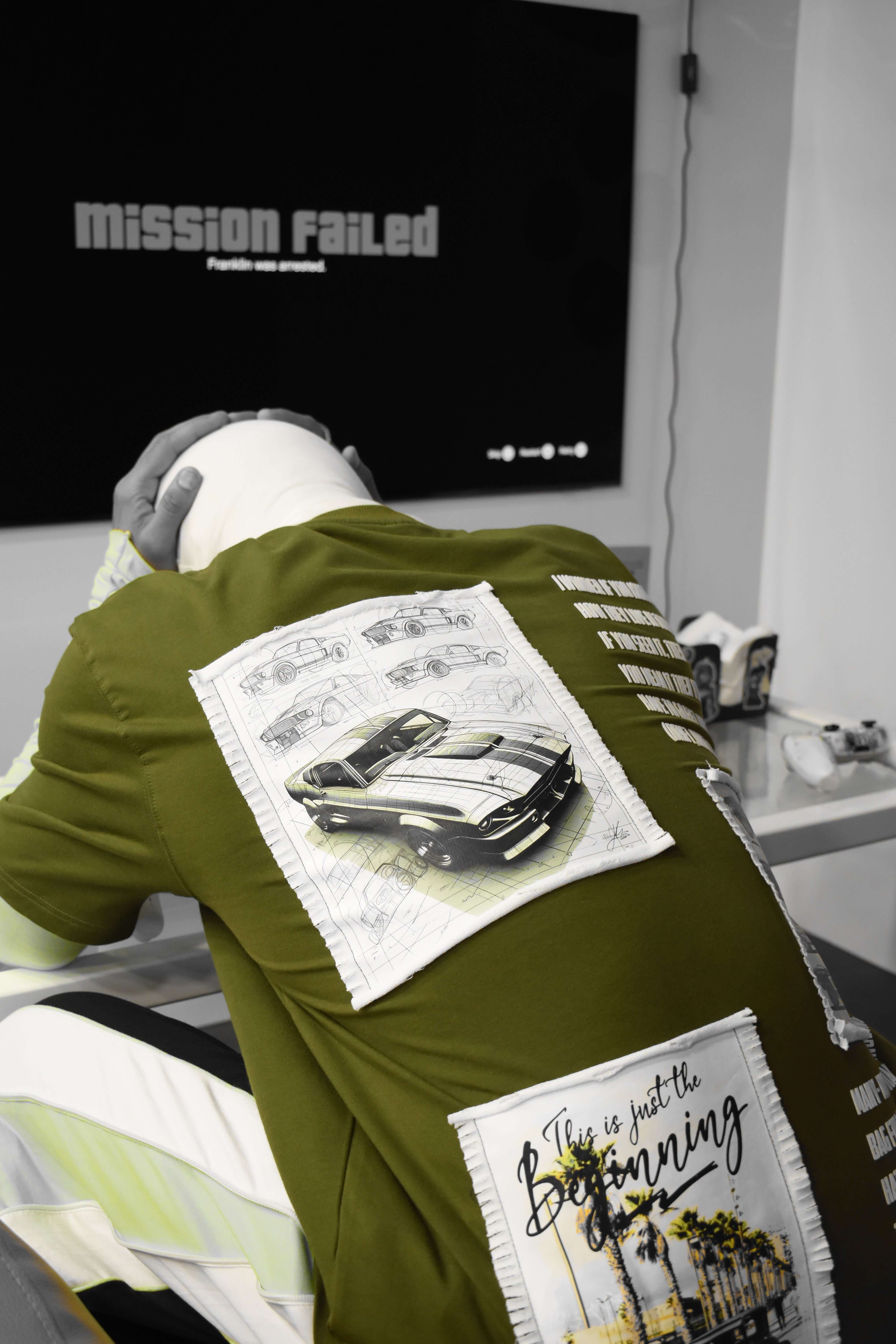 Olive green Ford Mustang designer over-sized T-shirt with car graphics, displayed on mannequin with video game console in background.