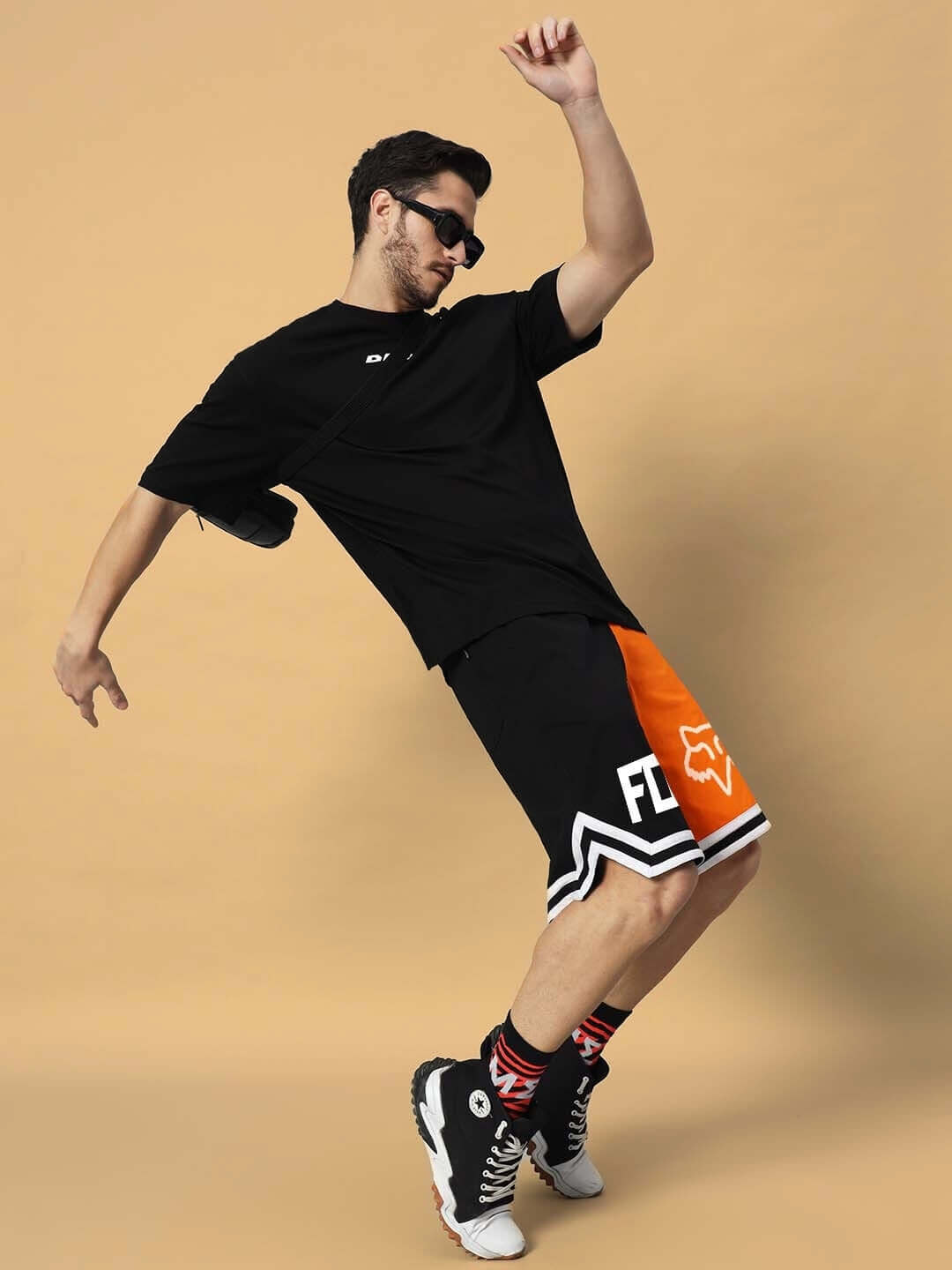 Man wearing FOX regular fit shorts in orange-black, styled with a black t-shirt and sneakers, showcasing dynamic movement against a neutral background.