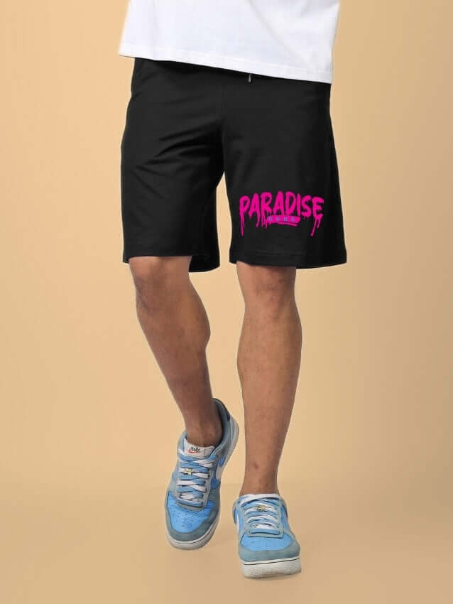 Person wearing FULNESS PARADISE SUMMER SHORTS BLACK with pink PARADISE text, paired with blue sneakers on beige background.