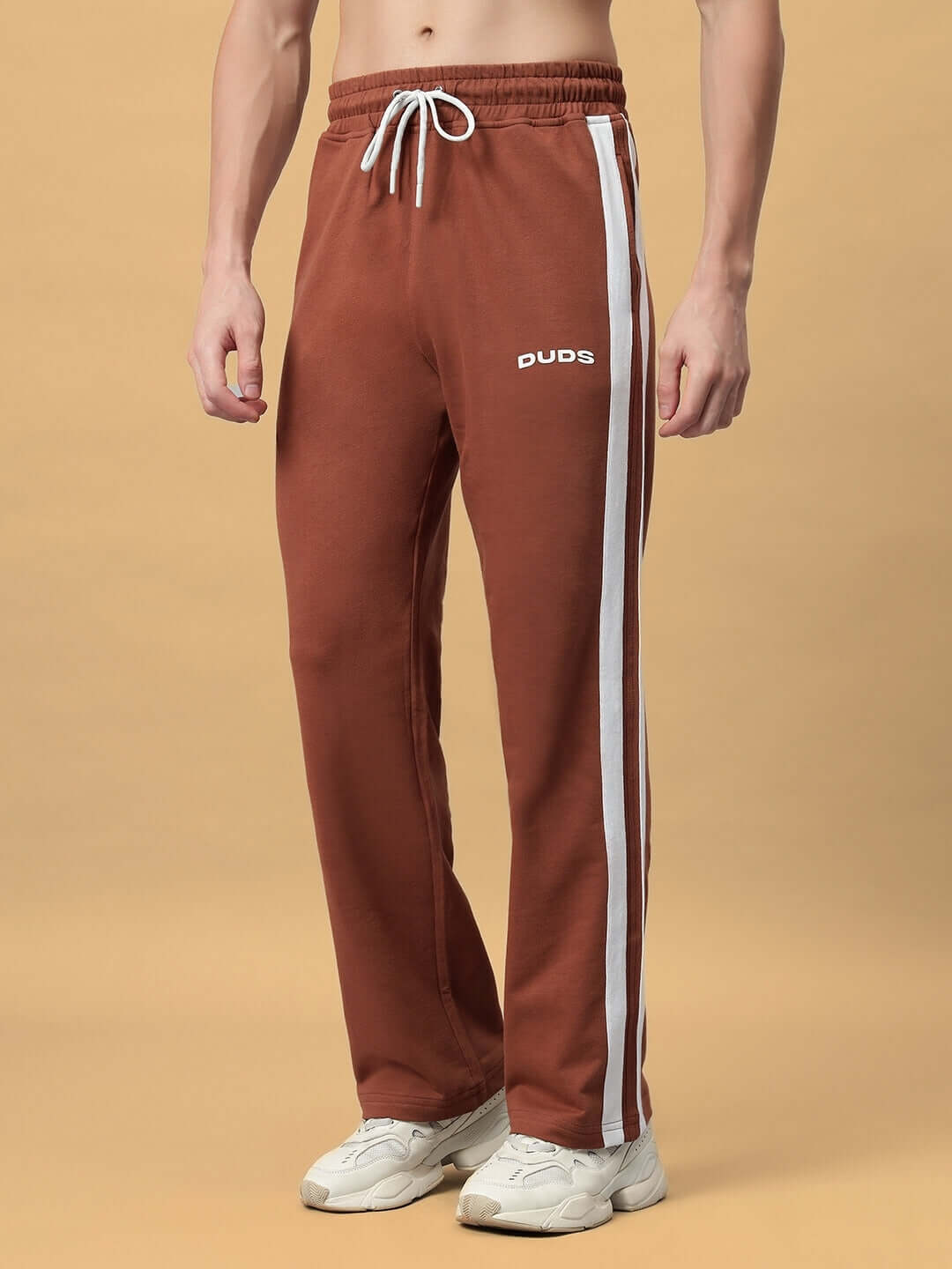 Model wearing FUTURE ICON 2 STRIPE JOGGER in brown with white stripes, crafted by DUDS, standing on beige background.