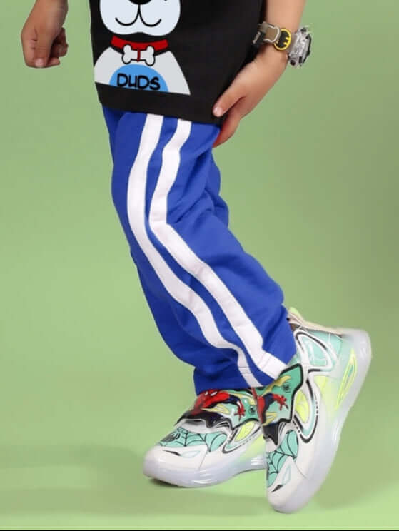Child wearing FUTURE ICON JOGGER FOR BOYS & GIRLS BLUE with white stripes, paired with colorful sneakers, over a green background.