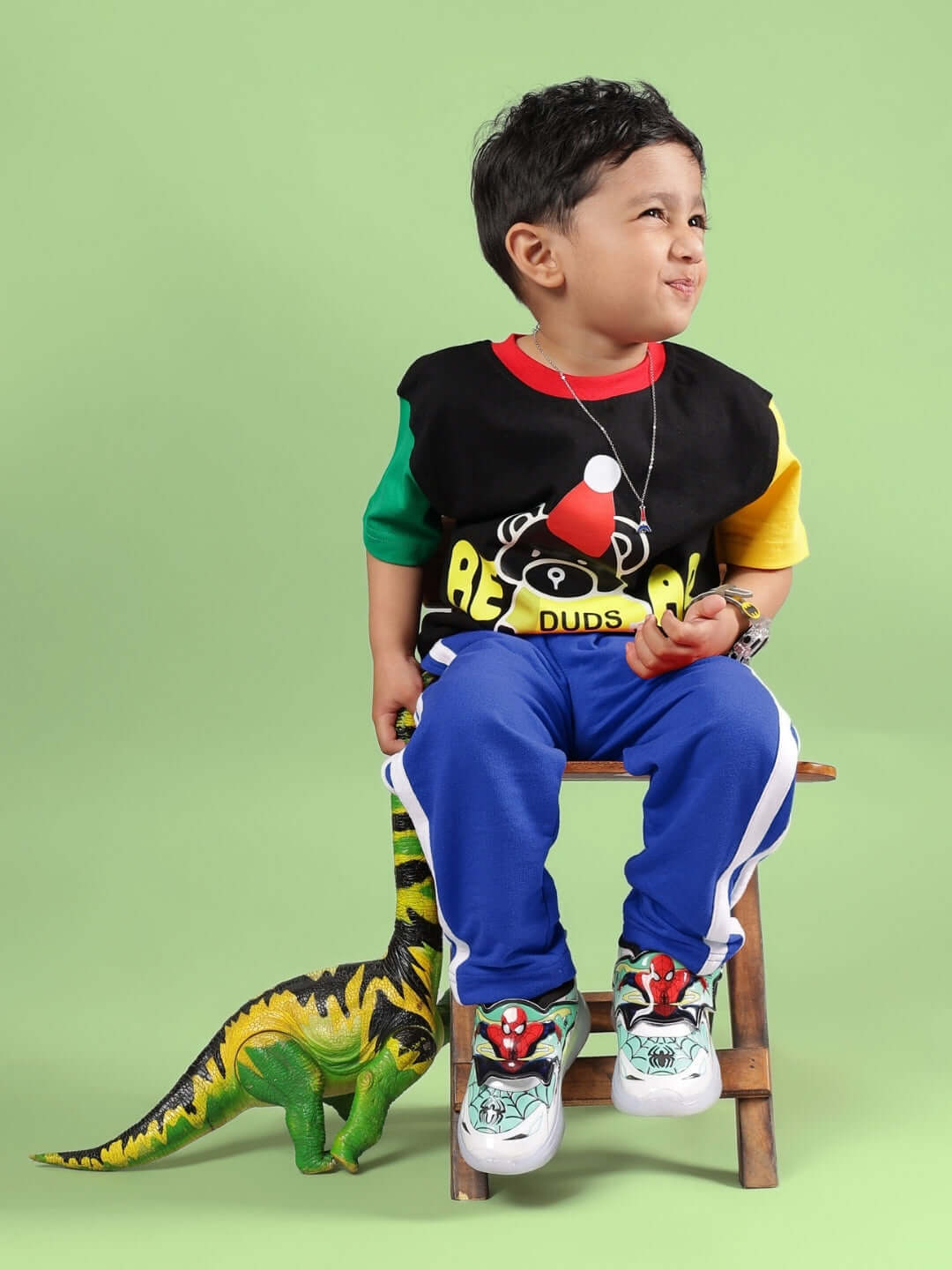 Child wearing FUTURE ICON JOGGER FOR BOYS & GIRLS BLUE with colorful shirt, sitting on a chair, green background, and toy dinosaur.