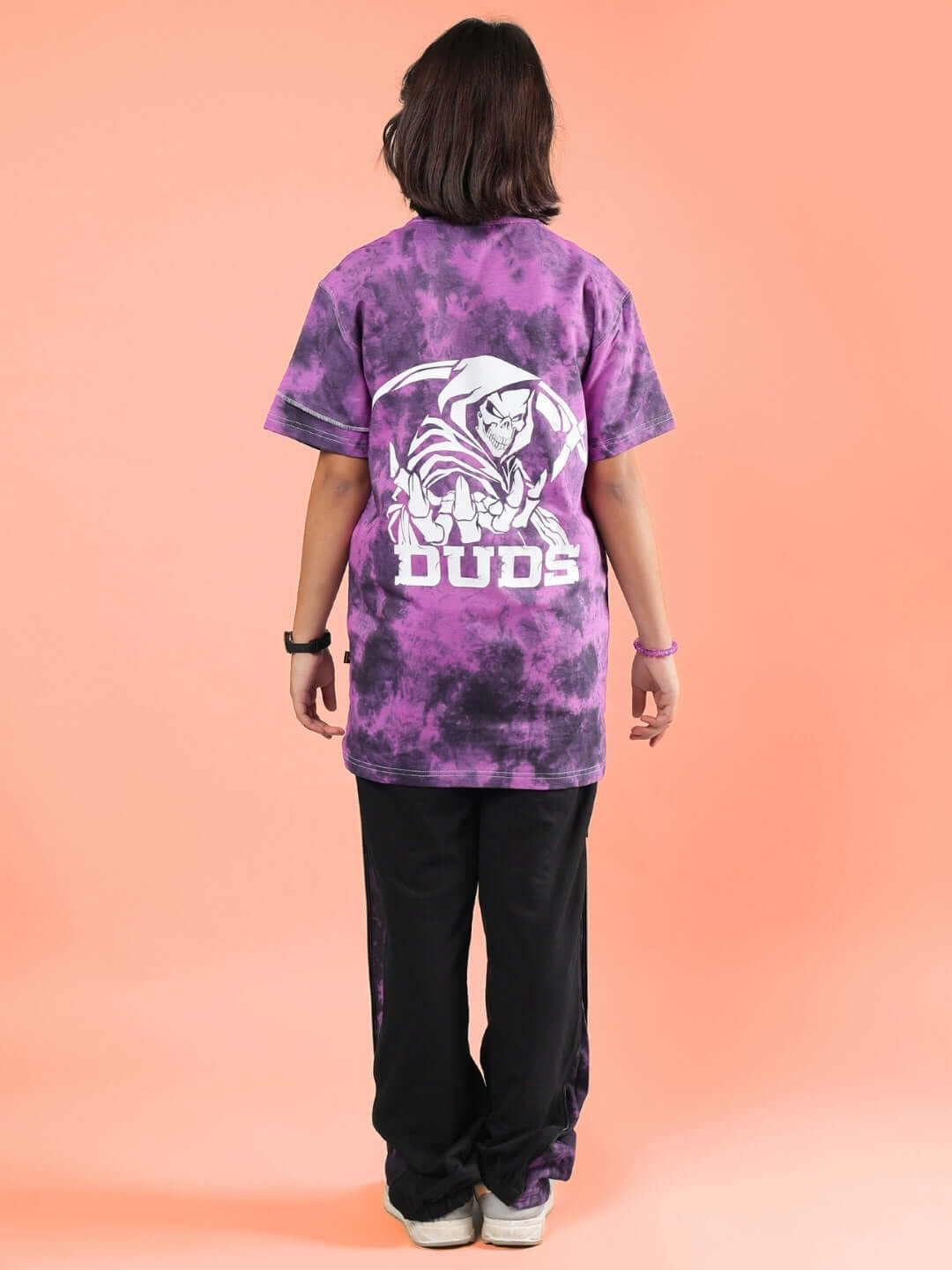 Child wearing GHOST RIDER CO-ORD FOR BOYS & GIRLS PURPLE BLACK outfit with DUDS logo on the back, standing against a peach background.