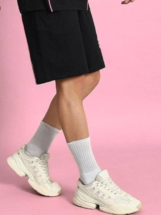 Person wearing GLARE REGULAR FIT SHORTS BLACK with white sneakers and socks on a pink background.