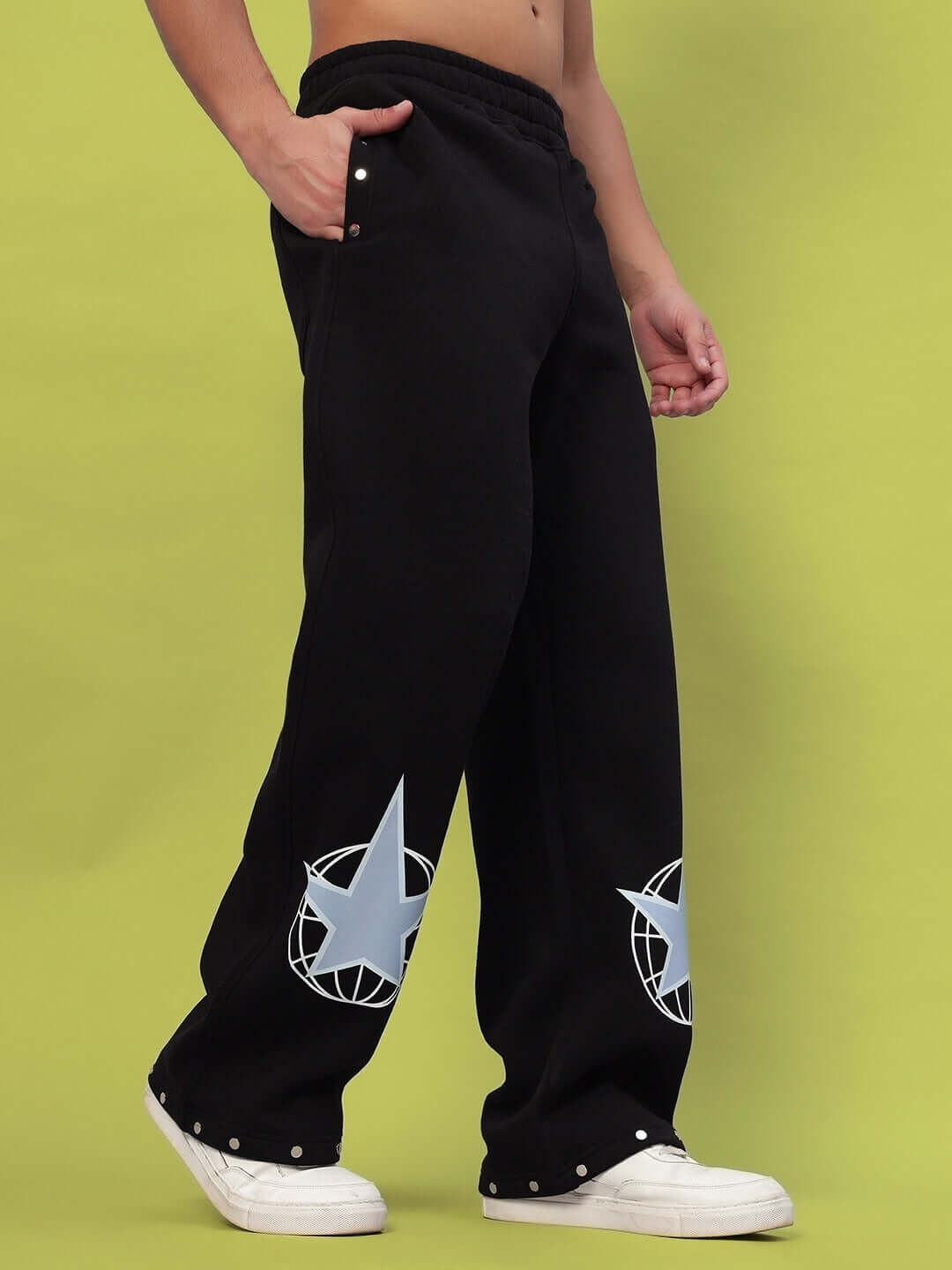 Black cotton fleece joggers with star prints, modeled with white sneakers on a green background. Ideal for casual wear.