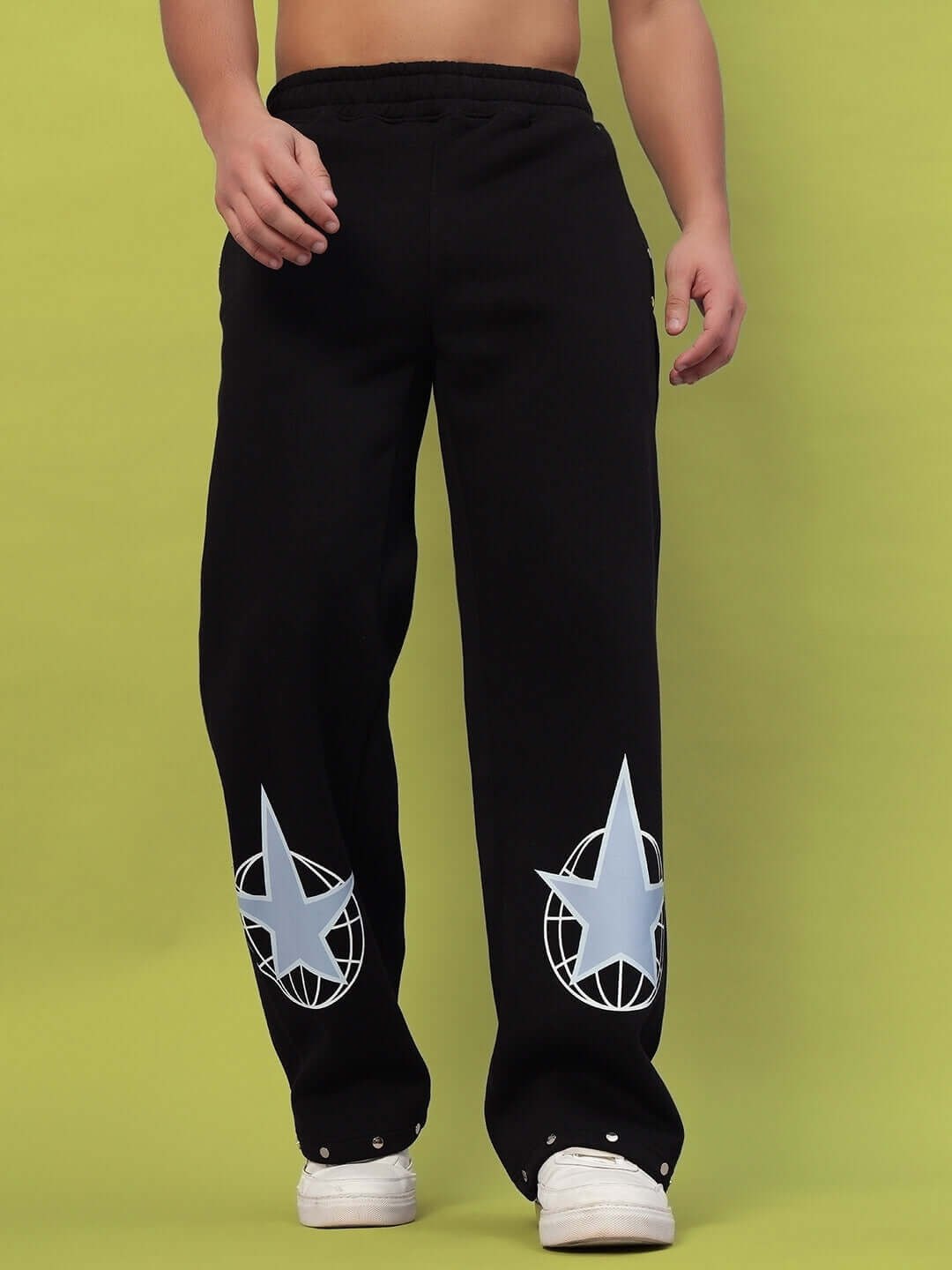 Man wearing GLOBAL COTTON FLEECE JOGGER BLACK pants featuring star graphics and elastic waistband, standing against green background.