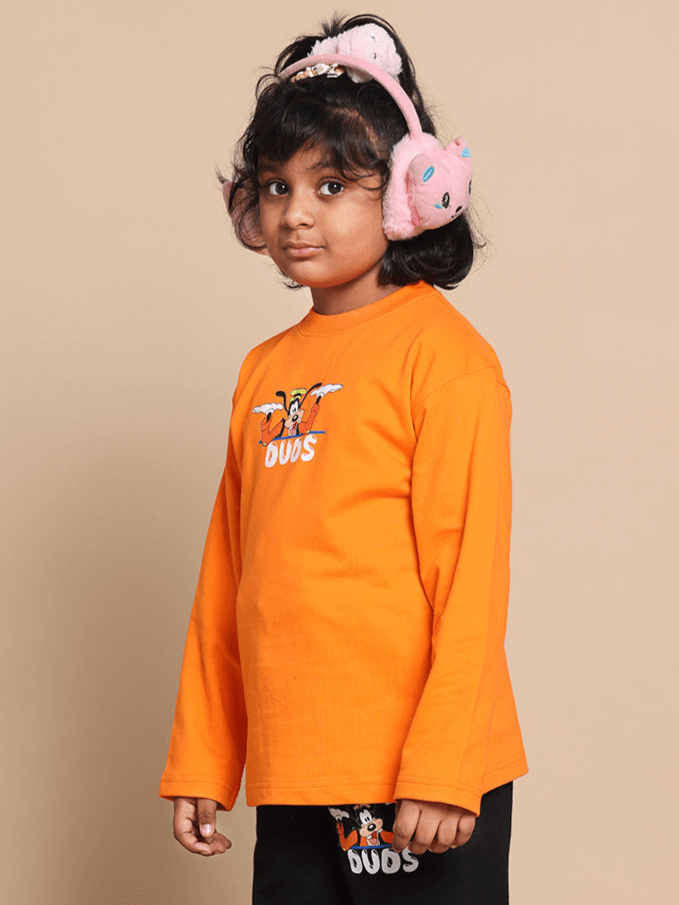 Child wearing an oversized orange sweatshirt with cartoon print, paired with pink earmuffs, standing against a beige background.