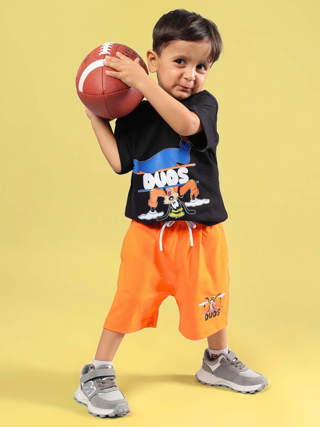 Child wearing GOOFY SUMMER CO-ORD FOR BOYS & GIRLS BLACK-ORANGE holding a football on yellow background.
