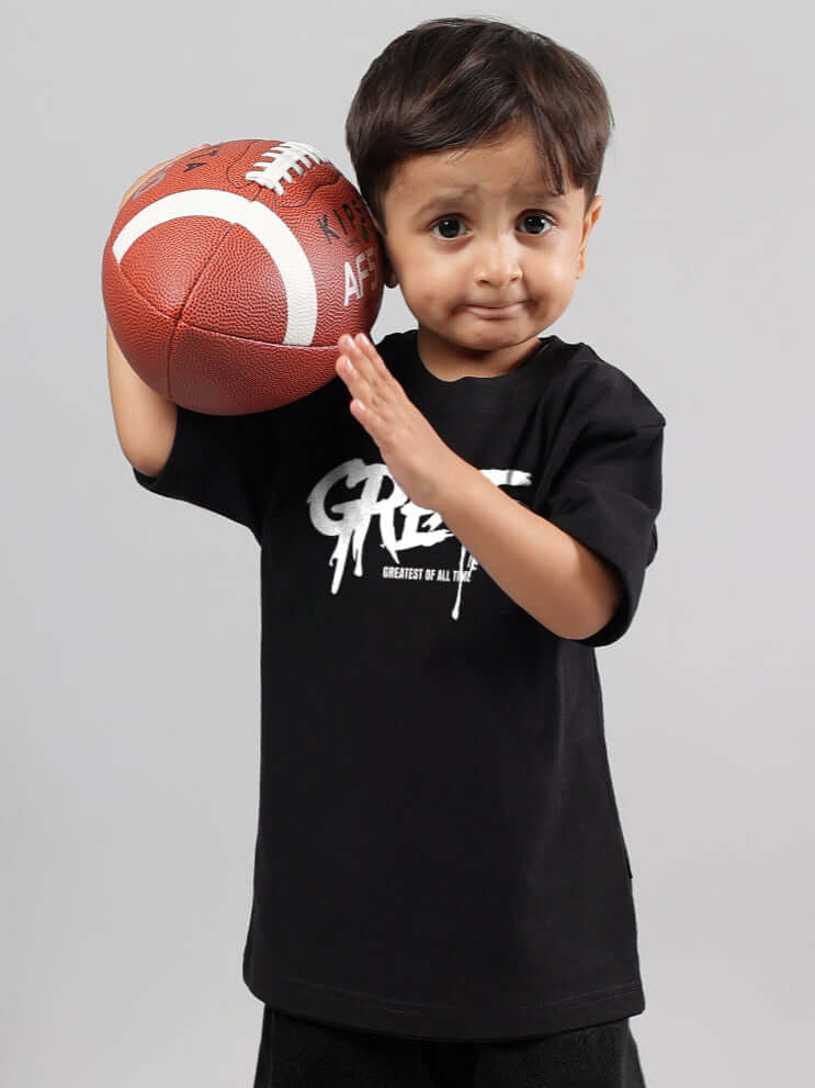Young child in black oversized t-shirt holding a football confidently.