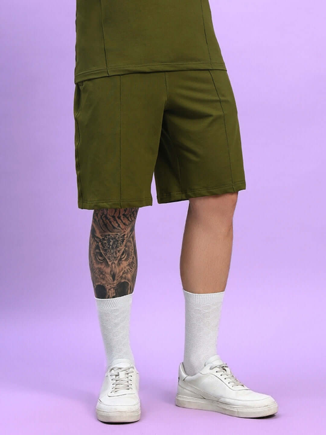 Olive green GREEBLE summer shorts, custom fit option, worn by a model with tattooed leg, white socks, and white sneakers on a purple background.