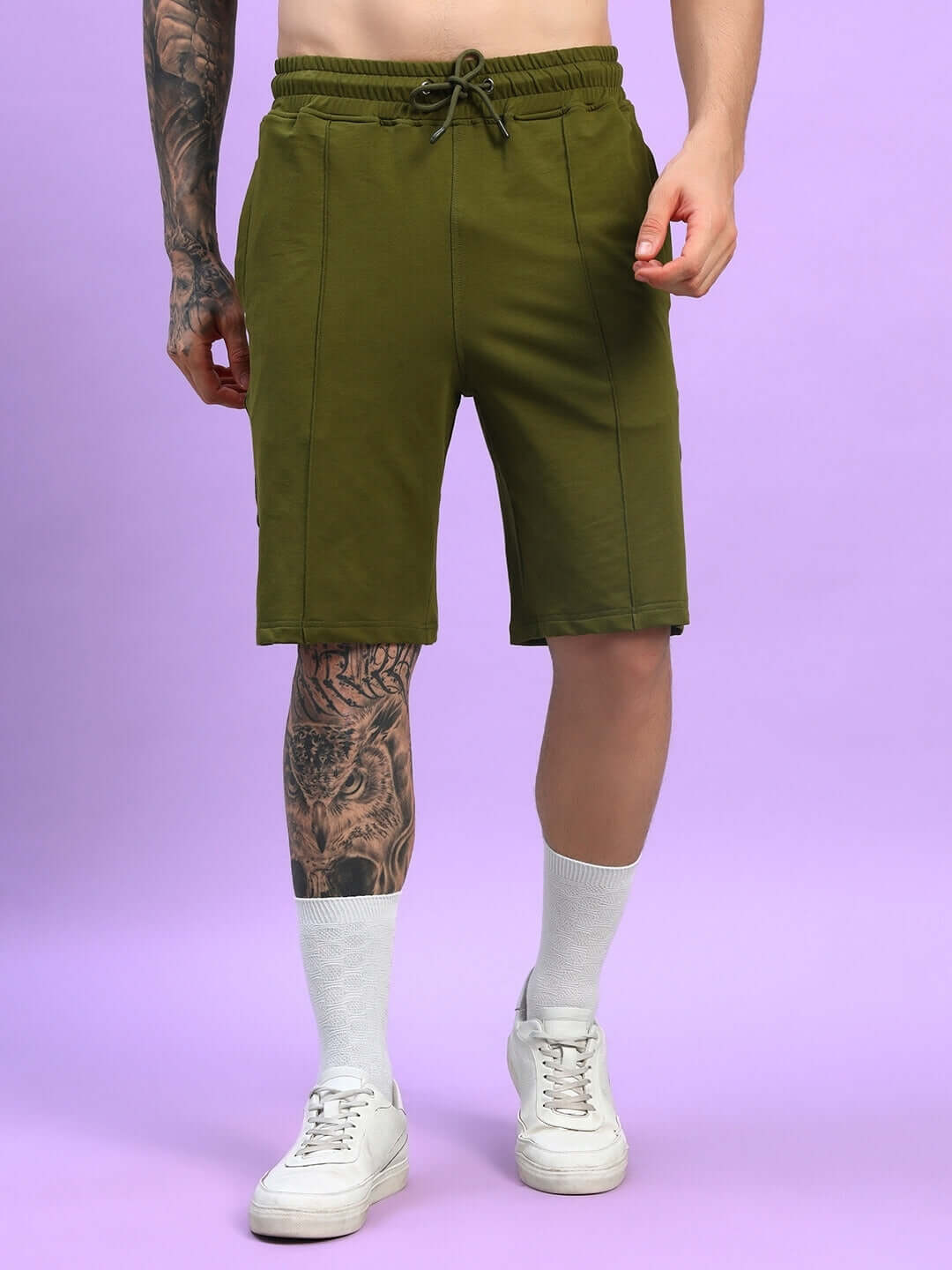 Man wearing olive green Greeble summer shorts, displaying front view and tattooed leg. Casual style with white sneakers and socks.