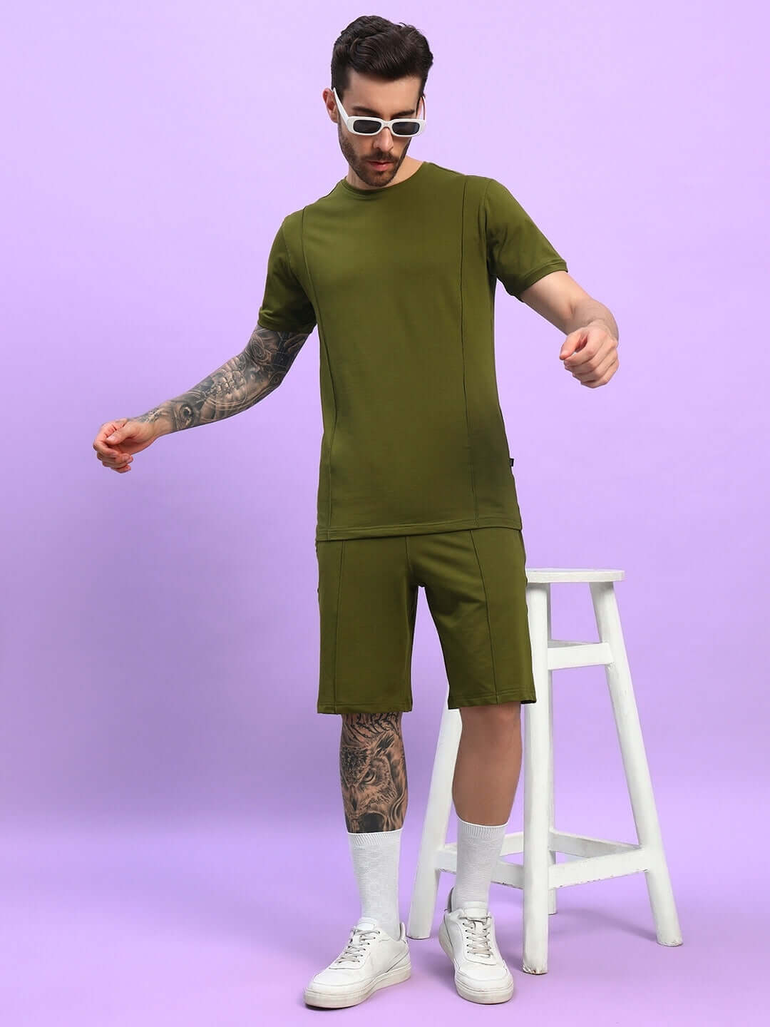 Model wearing GREEBLE Summer Shorts in Olive Green, paired with matching shirt, standing next to a white stool against a purple background.