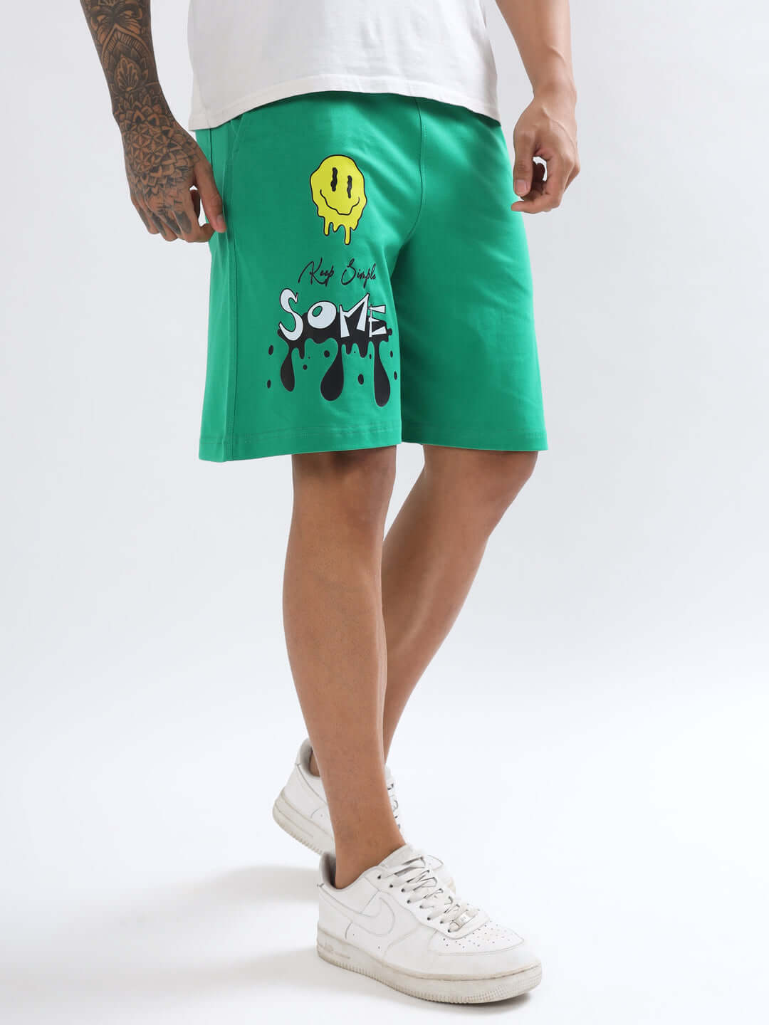 Man modeling green KEEP SIMPLE SOME shorts with smiley graphic, showcasing fit and style, wearing white sneakers on a neutral background.