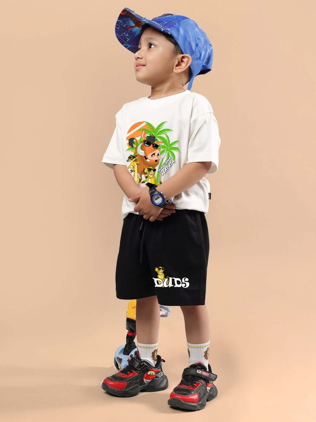 Child wearing DUDS Hakuna Matata Co-Ord set in off white and black with graphic print, blue cap, and colorful sneakers.