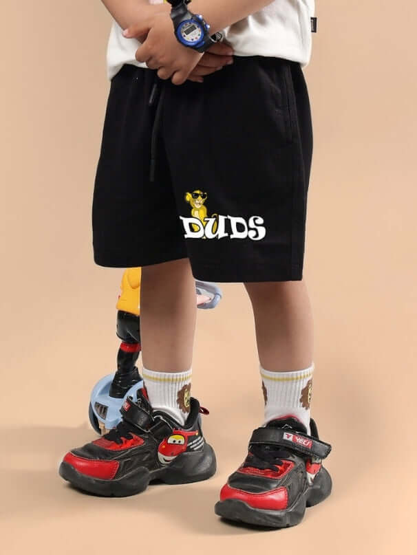 Child wearing HAKUNA MATATA SHORTS FOR BOYS & GIRLS BLACK, featuring 'DUDS' branding, styled with sporty shoes and socks, on a beige background.