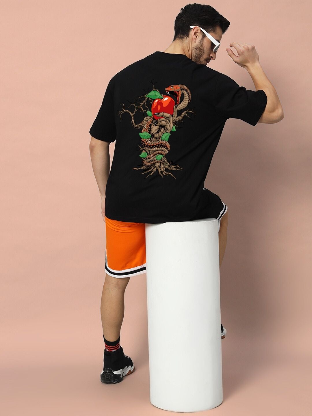 Model wearing HAND SNAKE APPLE OVERSIZED T-SHIRT BLACK with snake and apple design on the back, sitting on a stool.