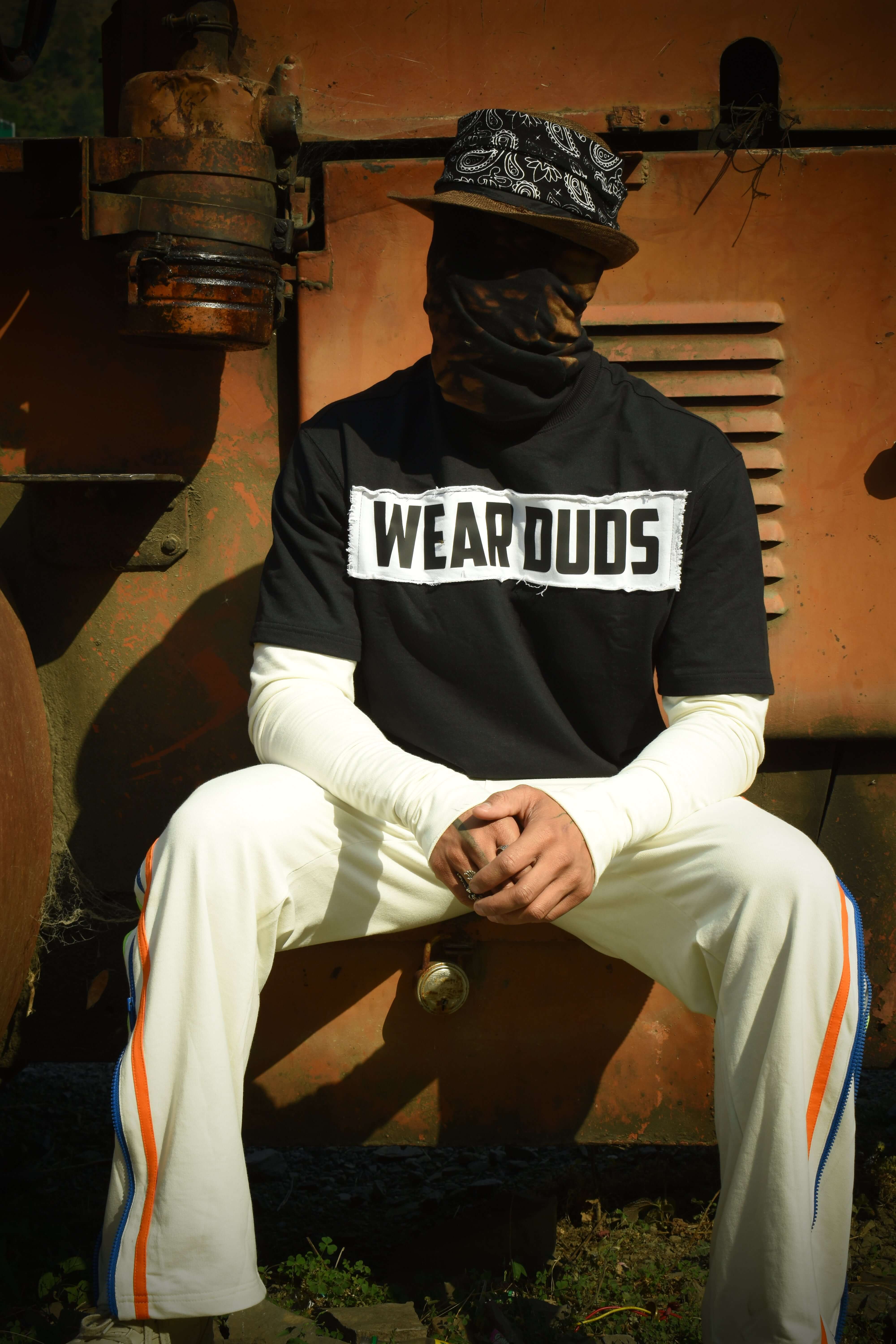 Man wearing a black WEAR DUDS t-shirt and white pants sitting in an urban setting with an industrial background.