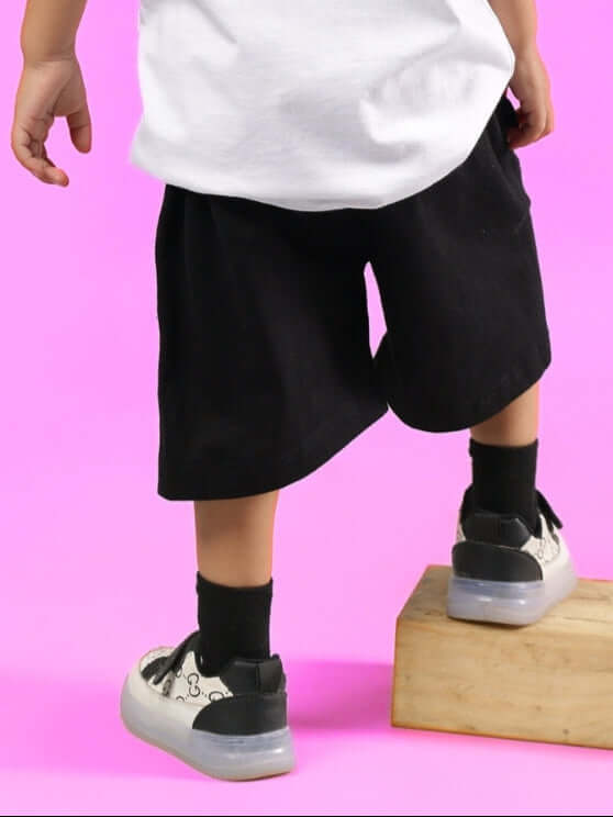 Child wearing black HEDGEHOG SHORTS FOR BOYS & GIRLS, standing on a wooden block against a pink background.