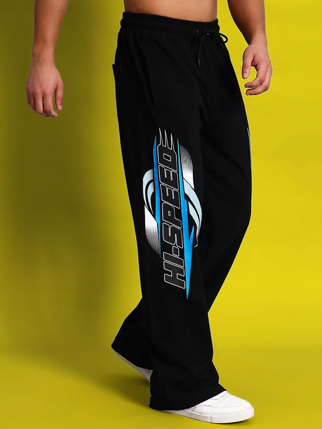 Man wearing black HI-SPEED relaxed fit jogger pants with white sneakers against yellow background.