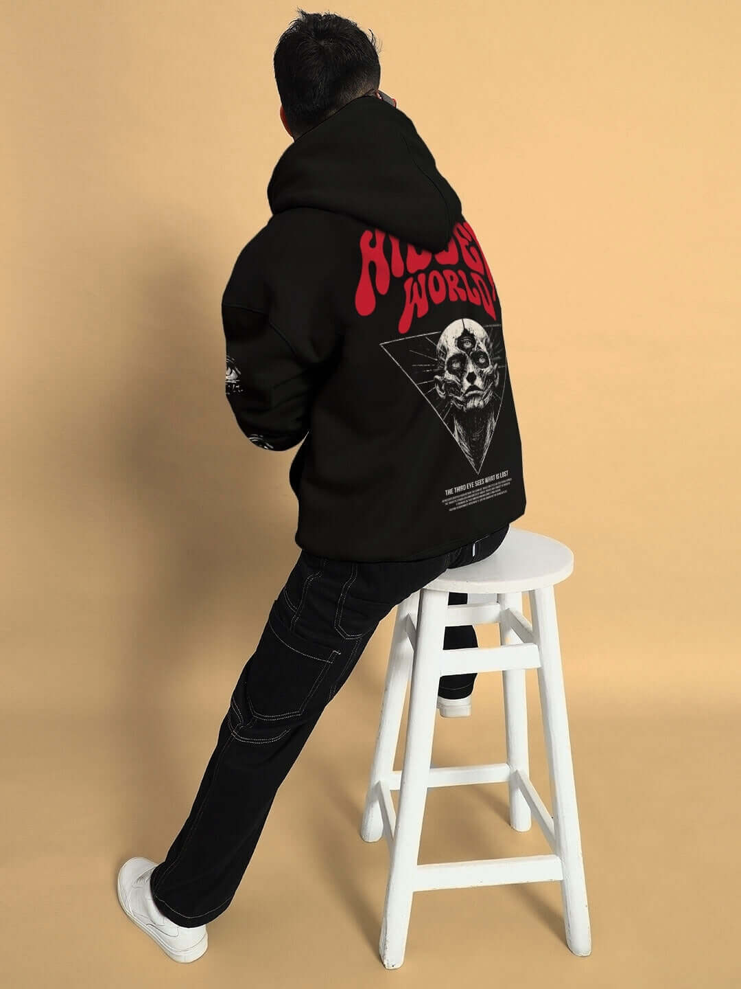 Person wearing Hidden World Oversized Fleece Hoodie in black, featuring bold graphic on the back, sitting on a stool against a beige background.