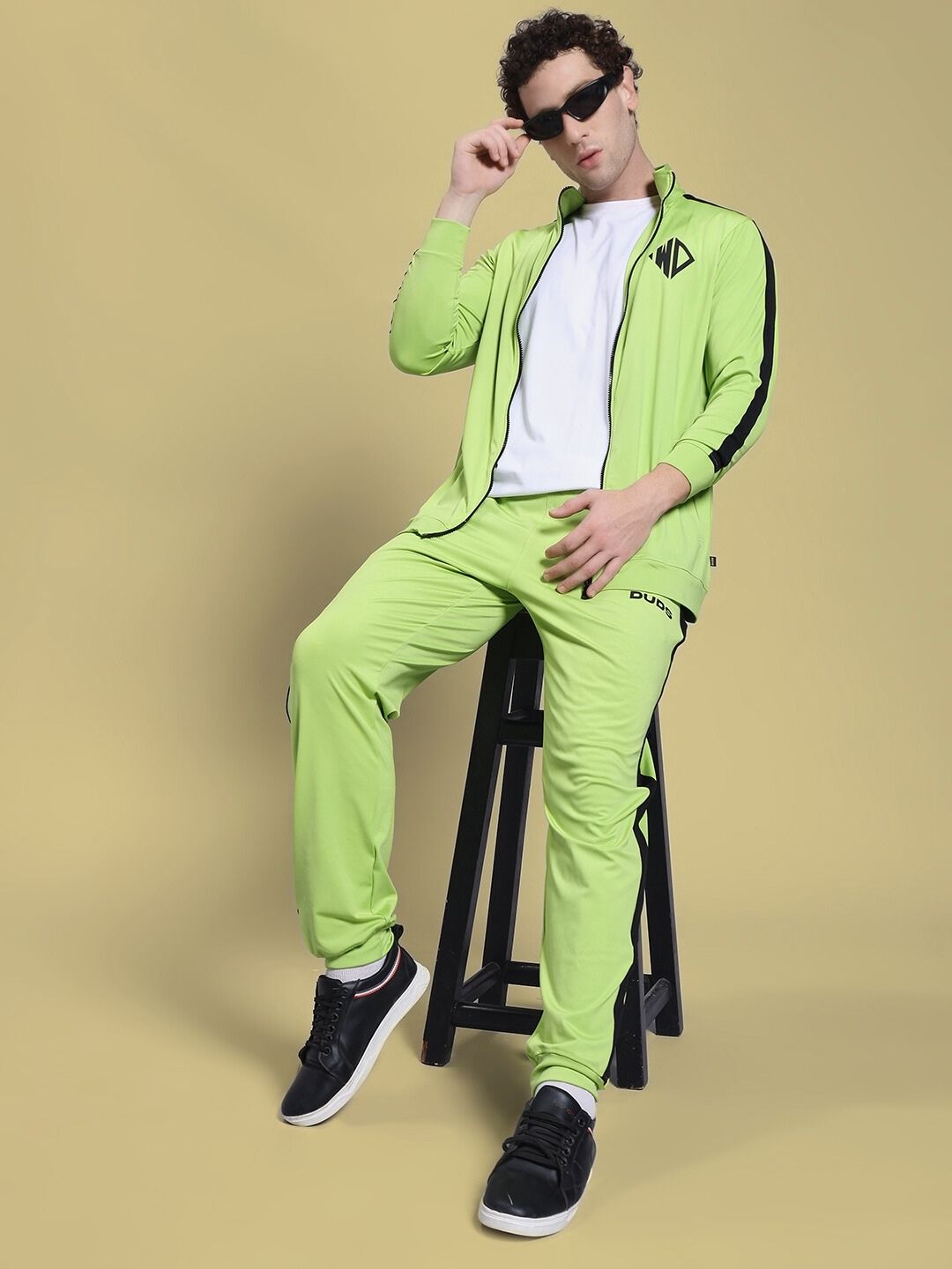 Model wearing HIGH PERFORMANCE JACKET NEON GREEN and matching pants, sitting on a stool.