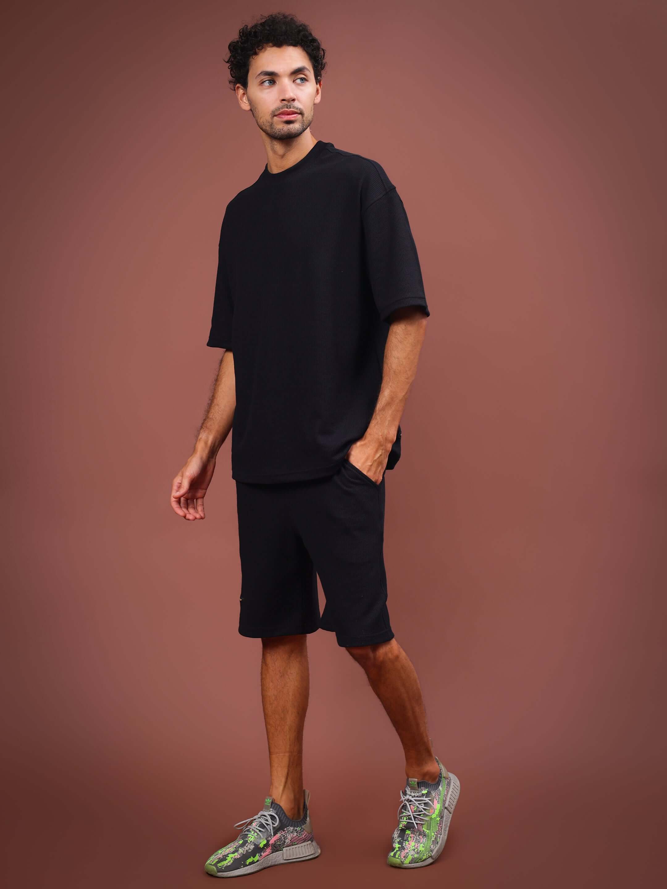 Man wearing HIGHKEY black solid co-ord set with casual sneakers, standing against a brown background.