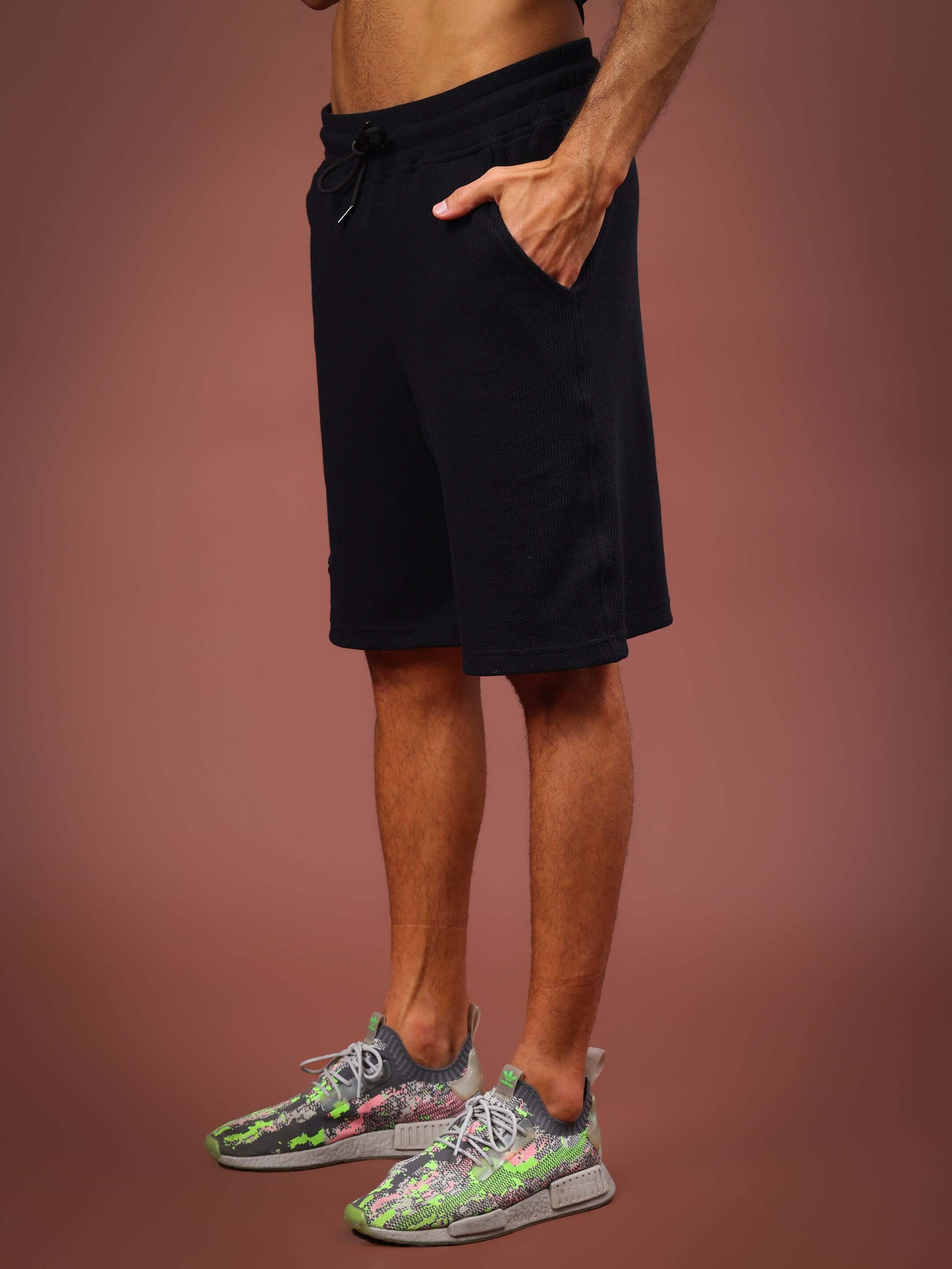 Male model wearing black HIGHKEY solid co-ord shorts, standing against a brown background, showcasing casual activewear.