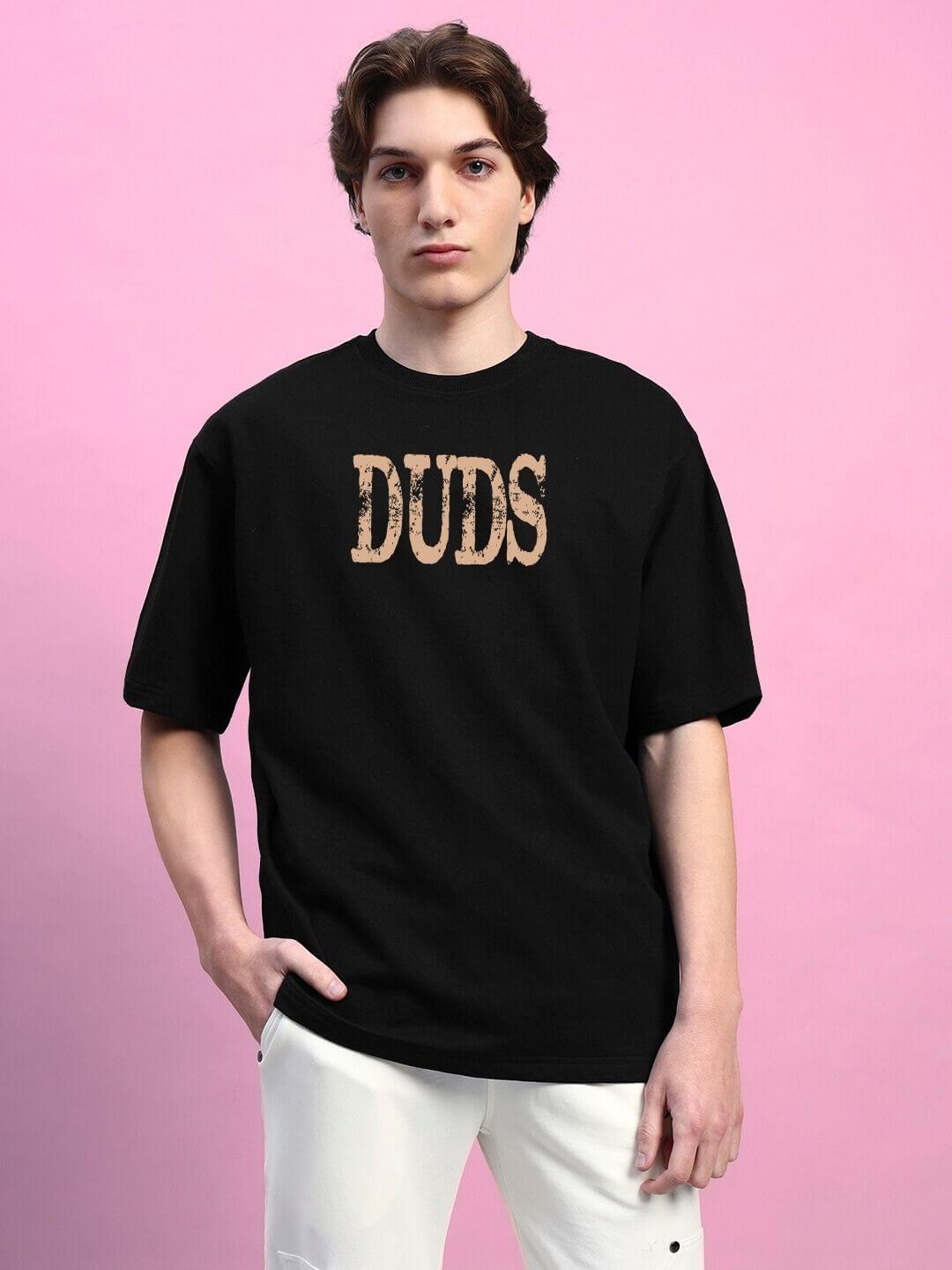 Model wearing a black oversized "DUDS" print T-shirt, premium cotton fabric, against a pink background.