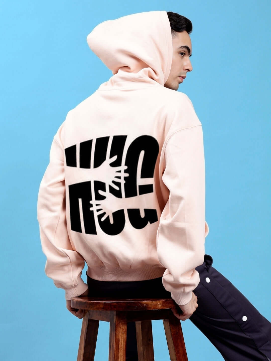 Person wearing HUG OVER-SIZED COTTON FLEECE HOODIES BEIGE, showing back design and relaxed fit, sitting on stool against blue backdrop.