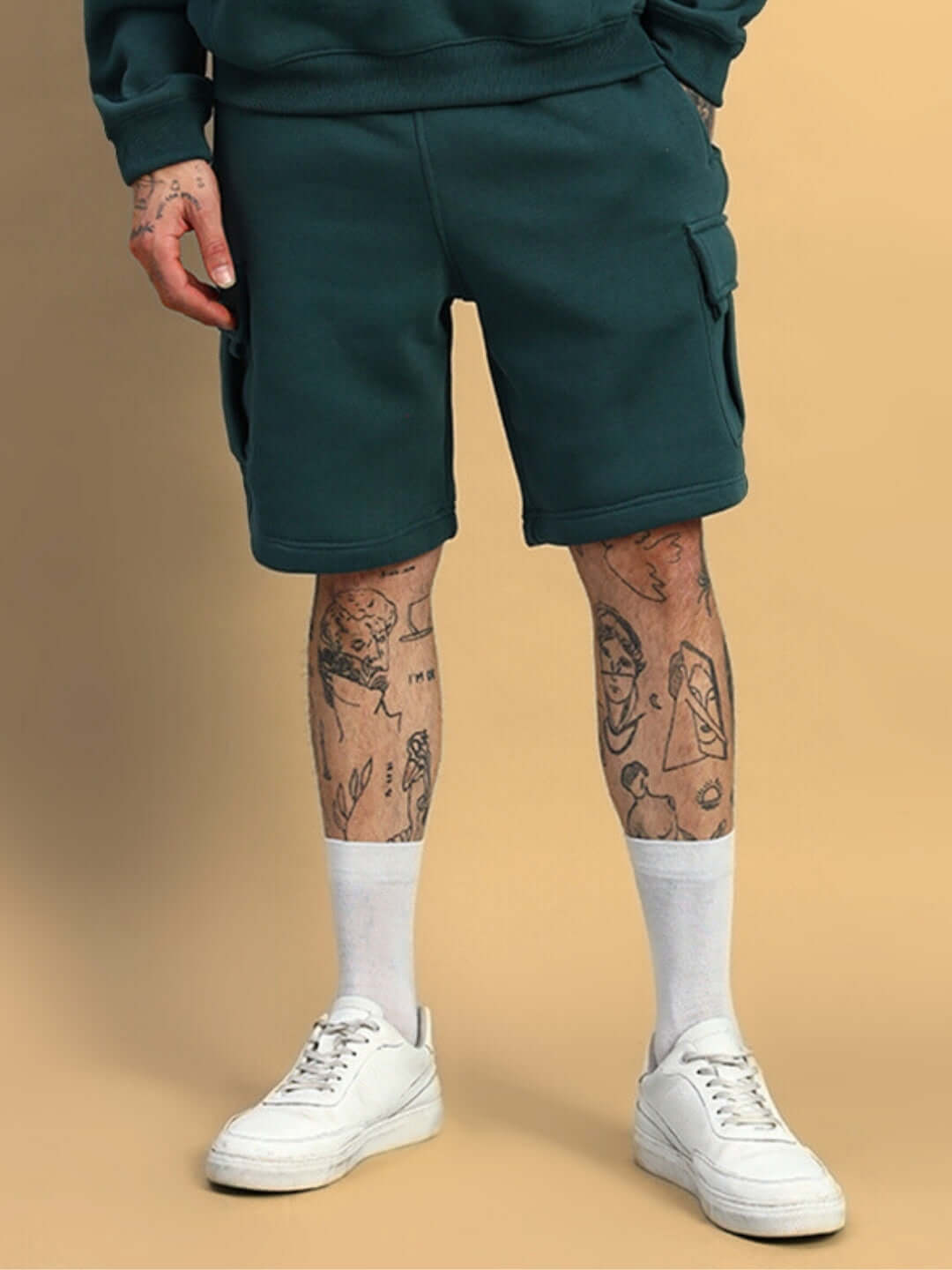 Man wearing HUSKY FLEECE SOLID CARGO SHORTS TEAL GREEN with tattoos and white sneakers, showcasing casual style.