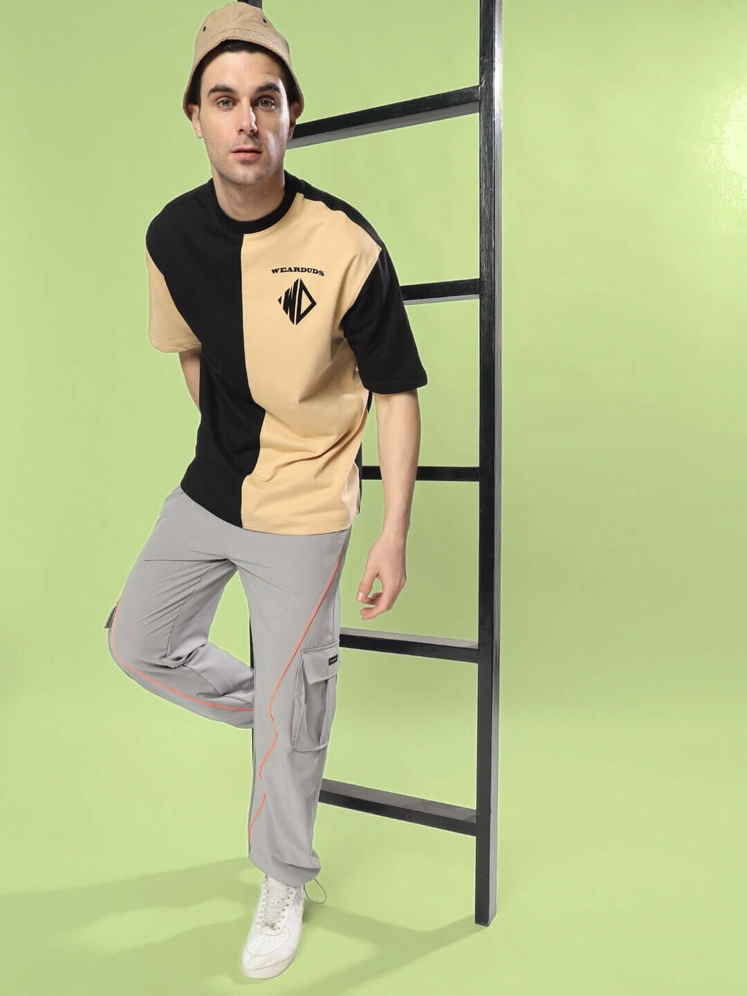 Model wearing HYPEBEAST grey cargo pants with neon orange accents and a stylish cap against a bright green backdrop.