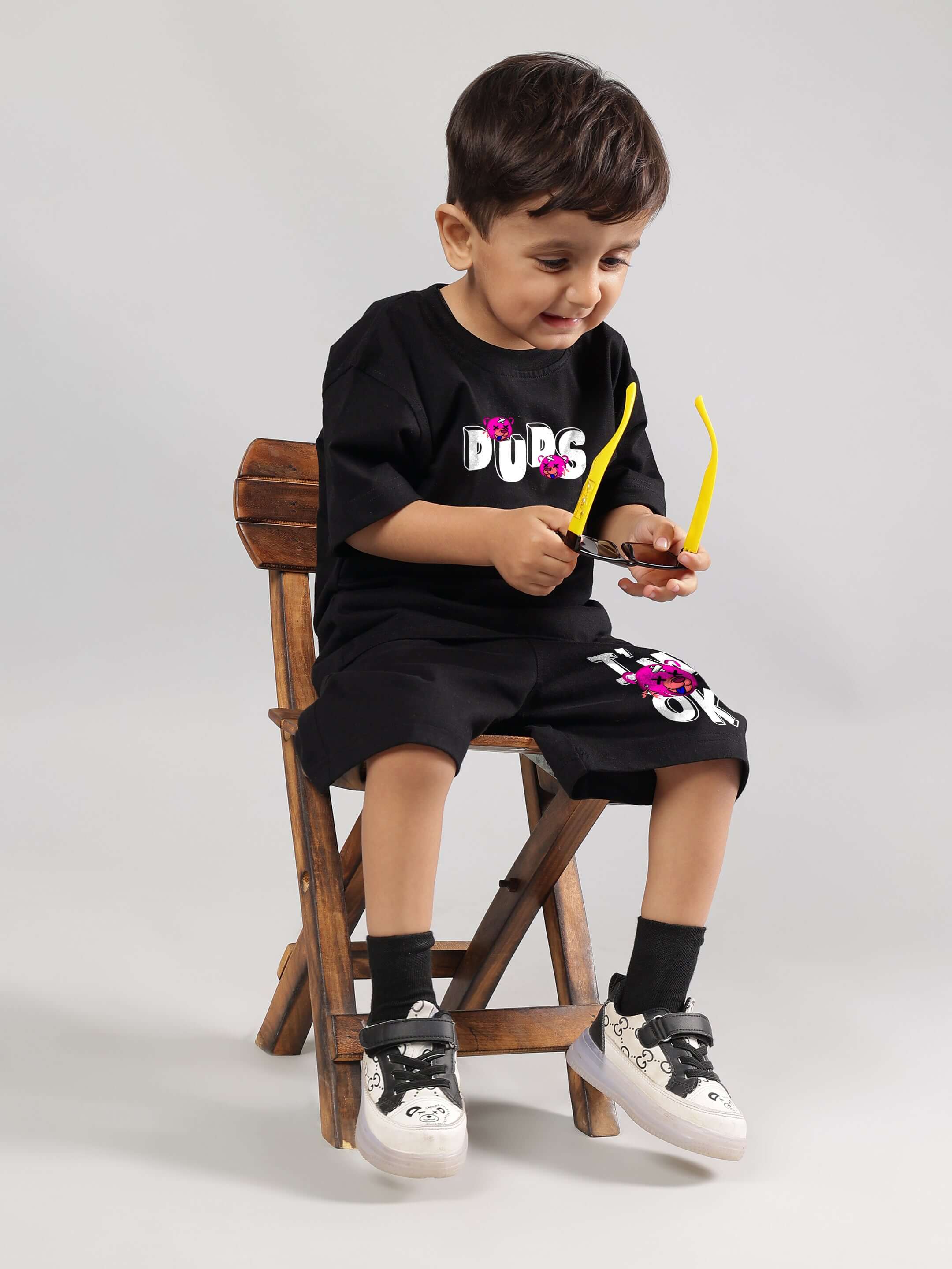 Boy wearing I'M OK Co-ord in black from DUDS, sitting on a wooden chair, holding yellow sunglasses, showcasing casual kidswear fashion.
