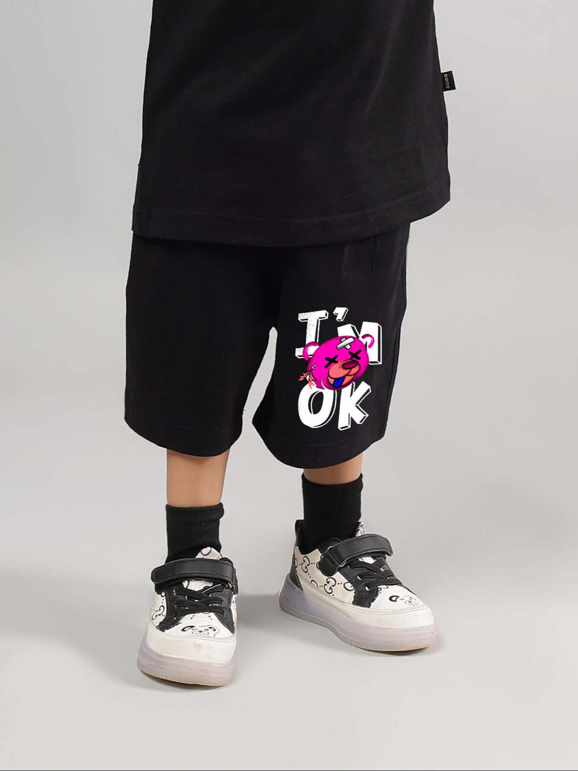 Child wearing I'M OK black shorts for boys and girls, featuring cartoon design, paired with black sneakers.