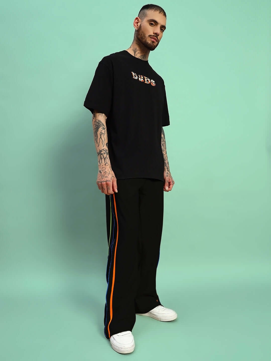 Man wearing black IMPERIAL SIDE ZIPPER JOGGERS by DUDS, showcasing side stripe design and relaxed fit, standing against mint green backdrop.