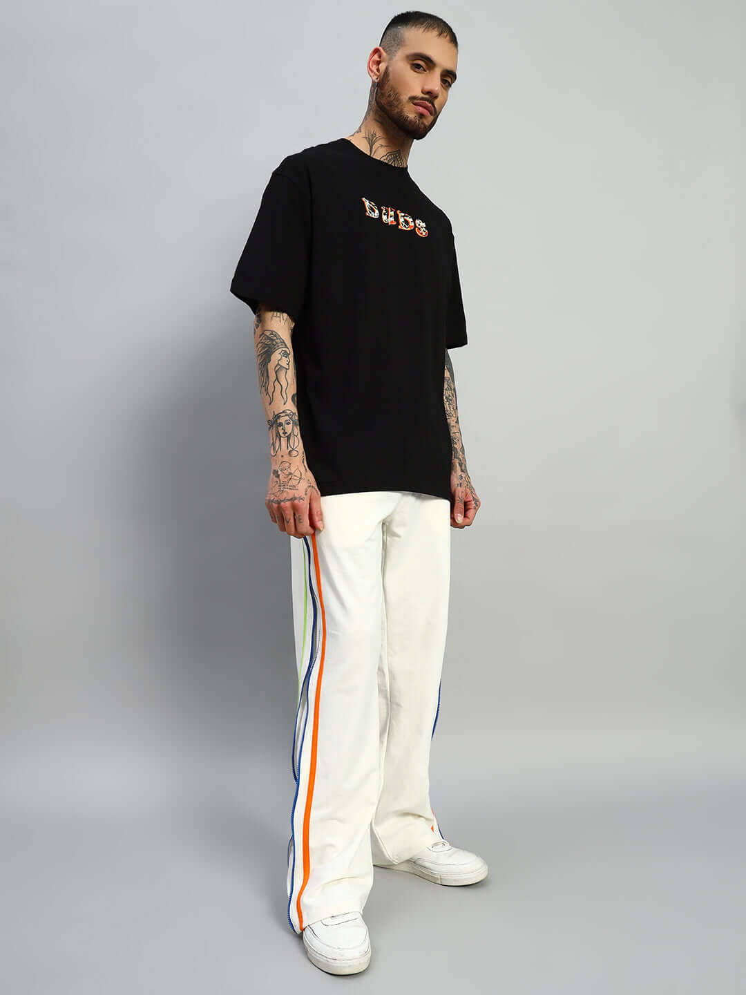 Model wearing Imperial Side Zipper Joggers in off-white with colorful side stripes, paired with a black DUDS t-shirt, standing on grey background.