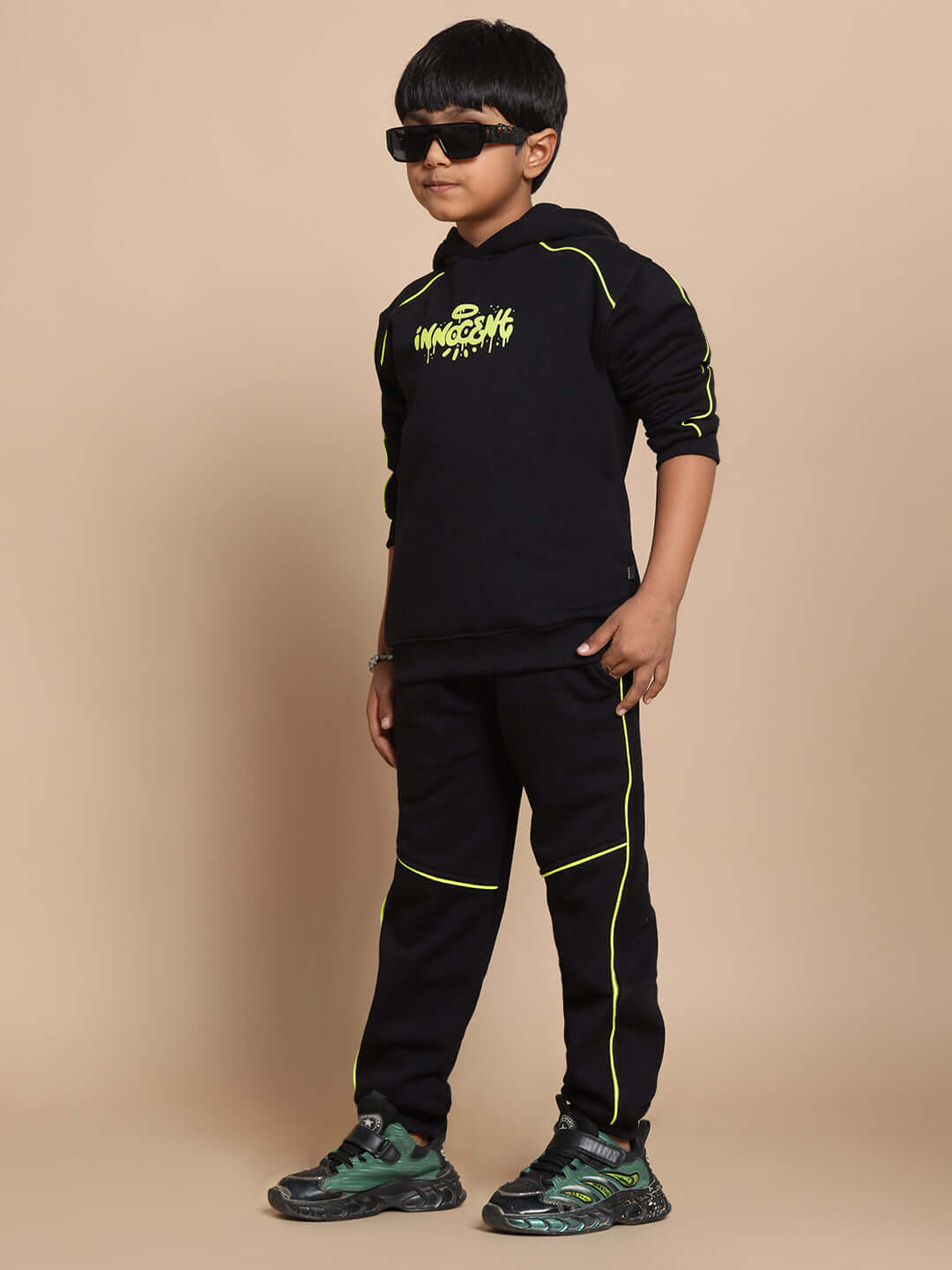 Young child wearing black INNOCENT jogging suit for boys and girls, with green accents, posing confidently with sunglasses.
