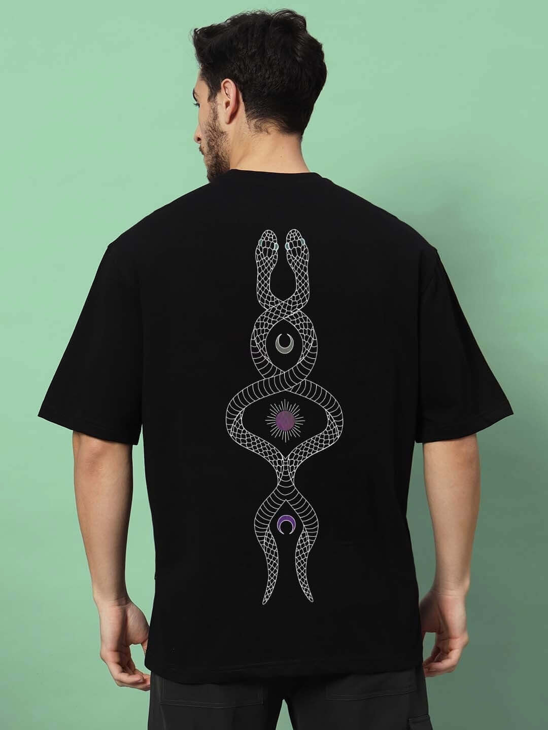 Man wearing black oversized t-shirt featuring intertwined snake reflector design on the back, standing against a mint green background.