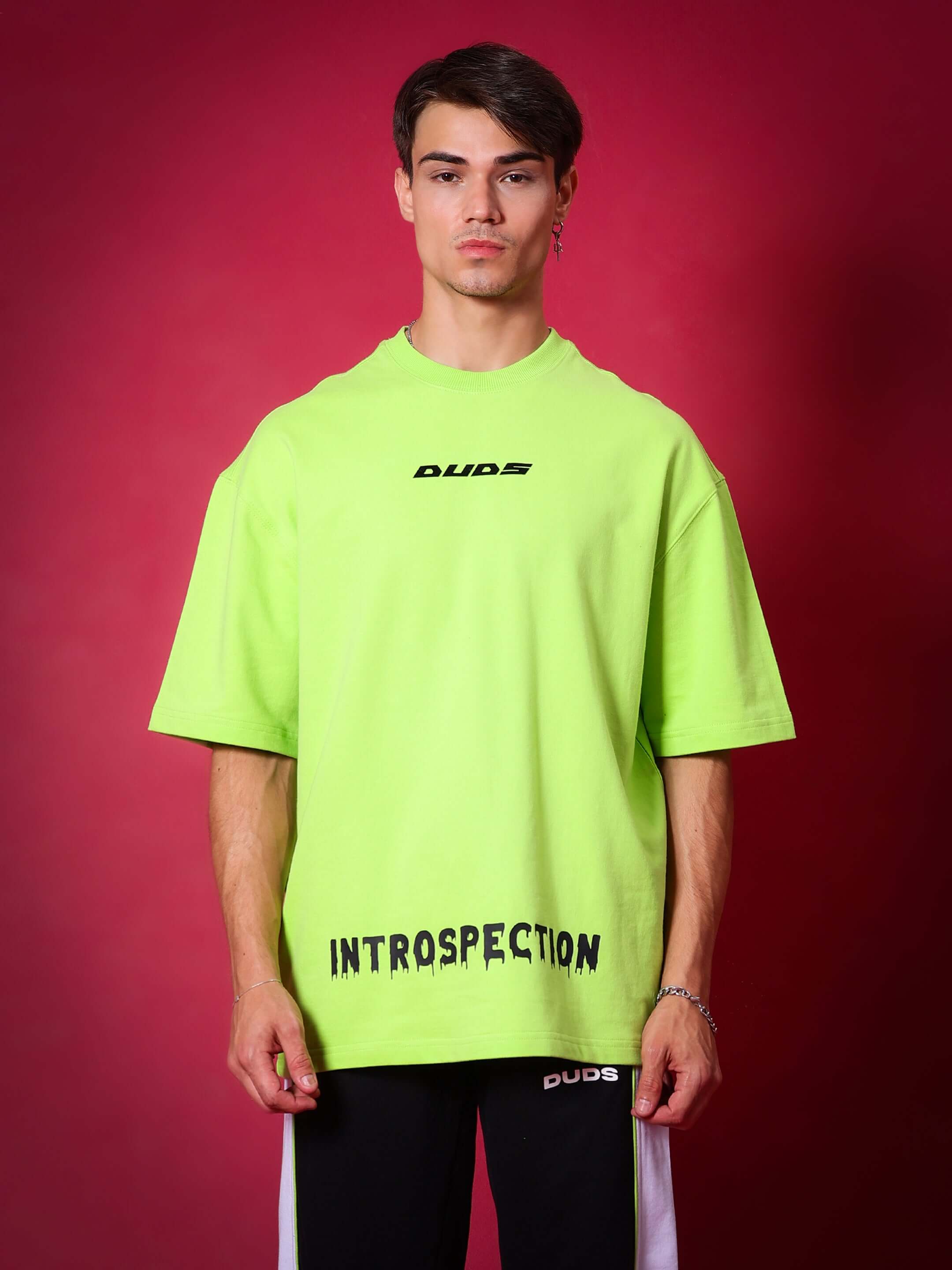 Neon green oversized t-shirt with 'Introspection' print, made of premium cotton blend, worn by a model against a red background.