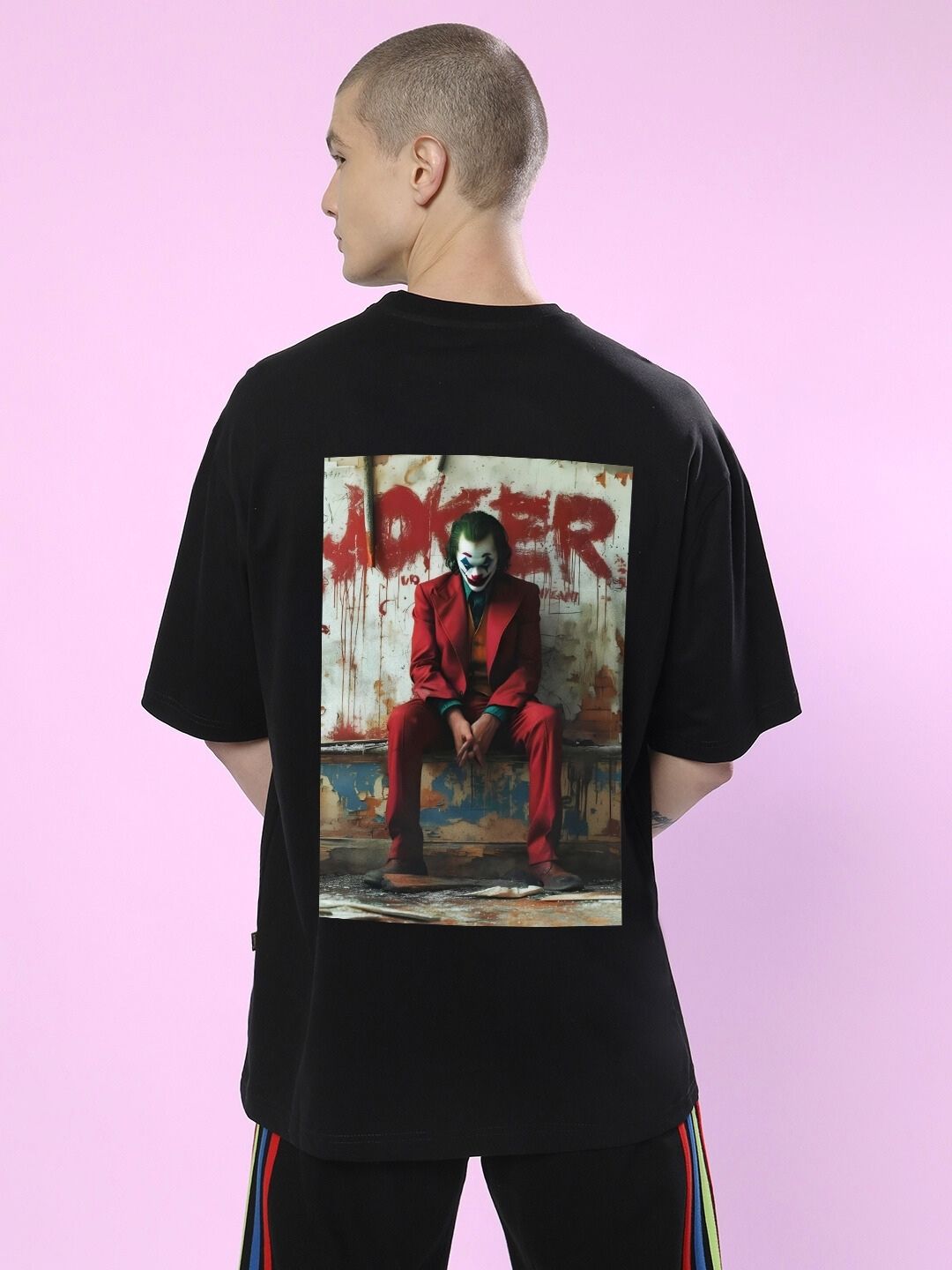 Man wearing JOKER PRINTED OVERSIZED T-SHIRT BLACK with graphic on the back, standing against a pink background.