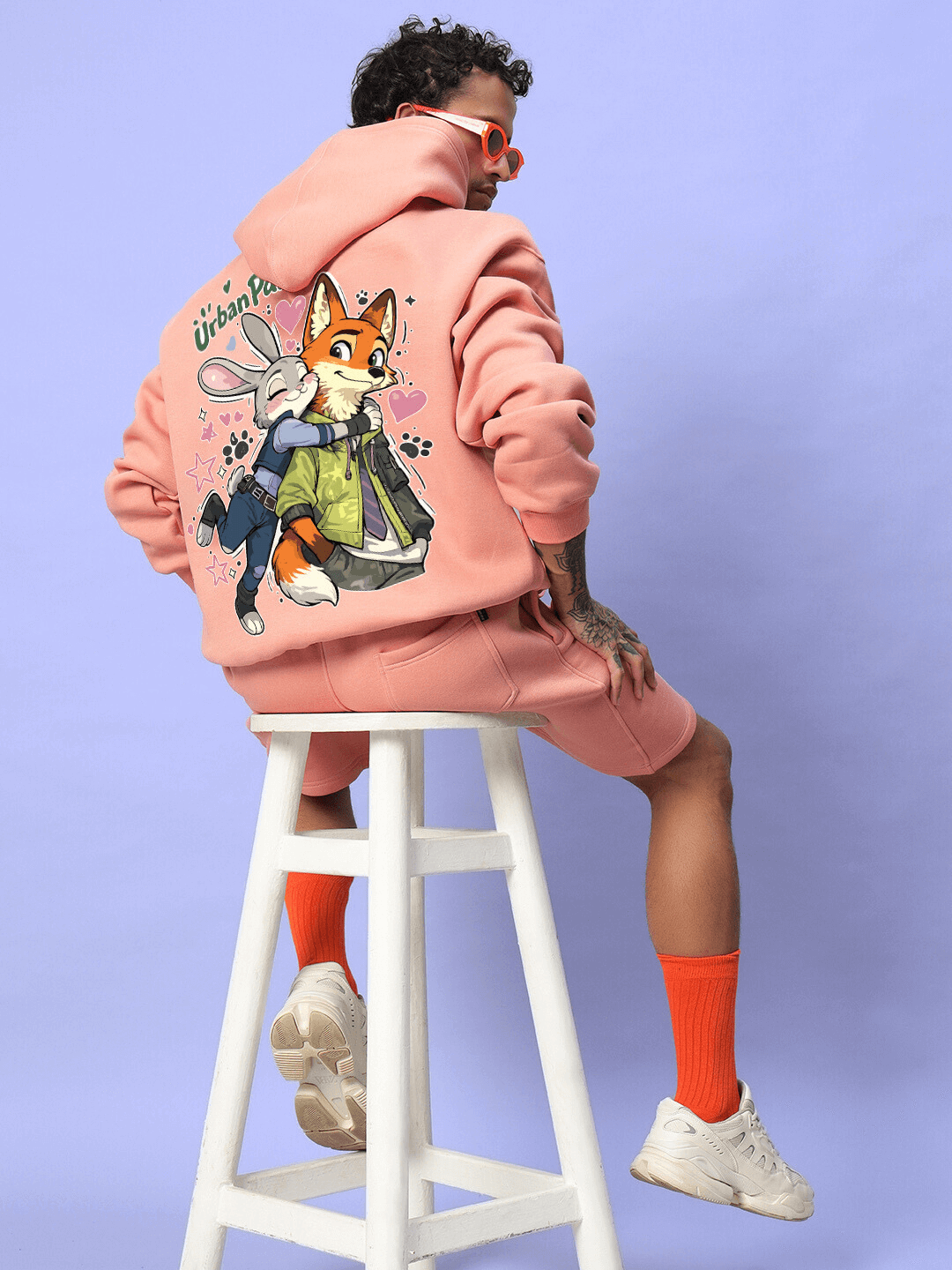 JUDY NICK ZOOTOPIA FLEECE HOODIE PEACH featuring characters from Zootopia, worn by a model sitting on a stool against a purple background.