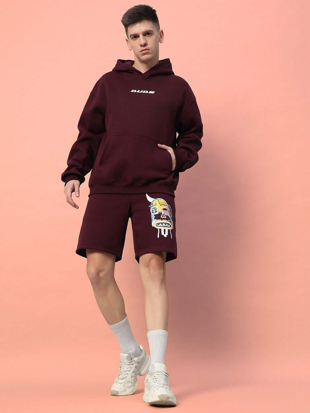 Model wearing JUMBLED FLEECE CO-ORD WINE with graphic print, featuring a hoodie and shorts, against a peach background.