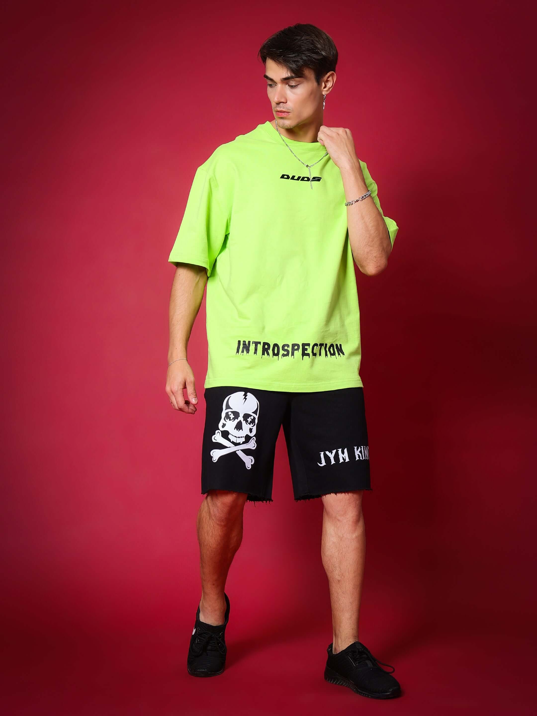 Model wearing JYM KING Regular Fit Black Shorts with a skull print on the left side, paired with a neon green t-shirt, on a red background.