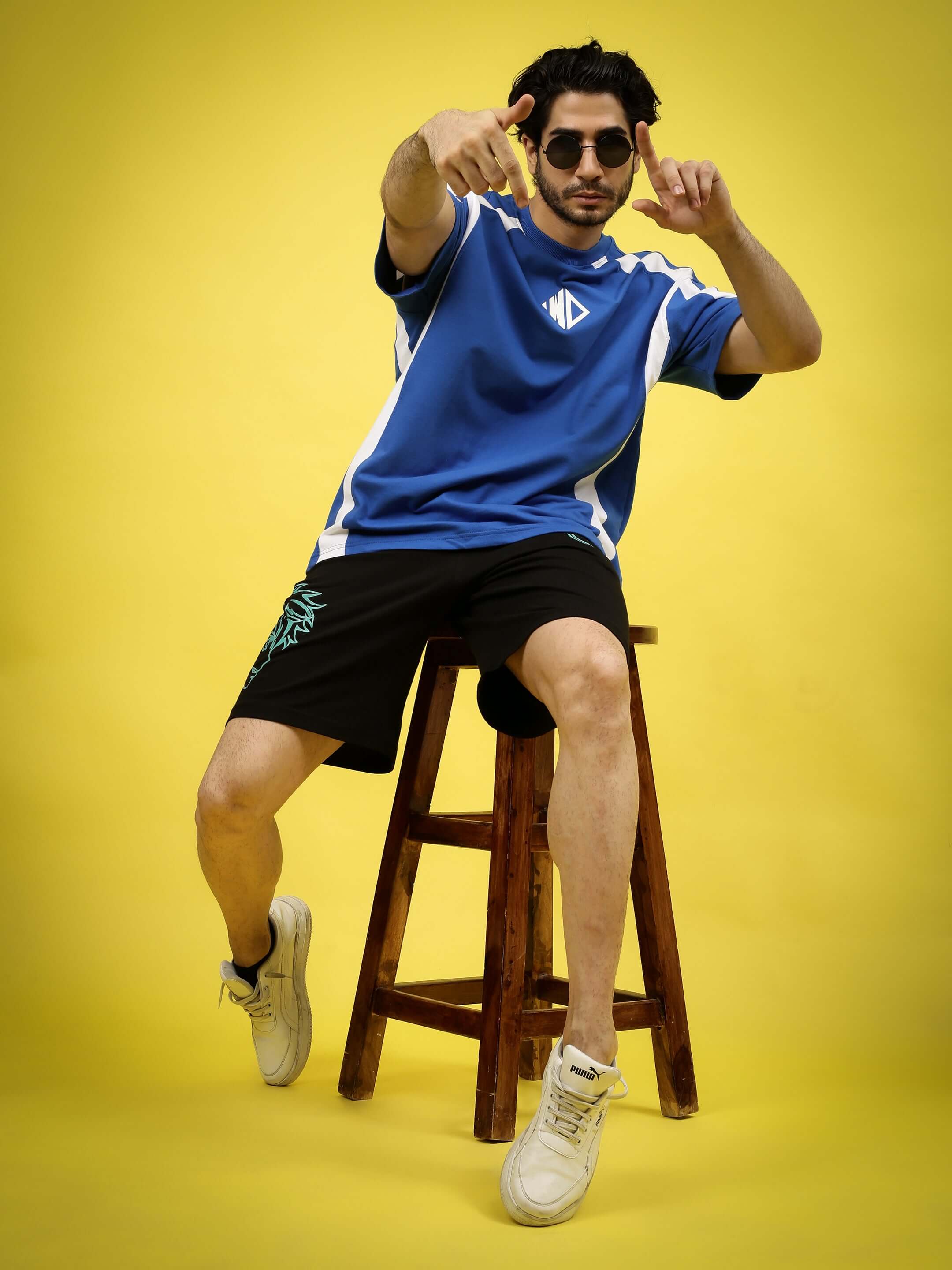 Model wearing blue jersey and black Kakashi regular fit shorts, sitting on a stool with a yellow background.